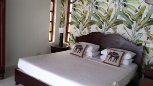 Un lit avec une literie blanche et deux coussins décoratifs, une tête de lit en bois, une table de chevet avec une lampe et une fenêtre avec des stores verticaux.