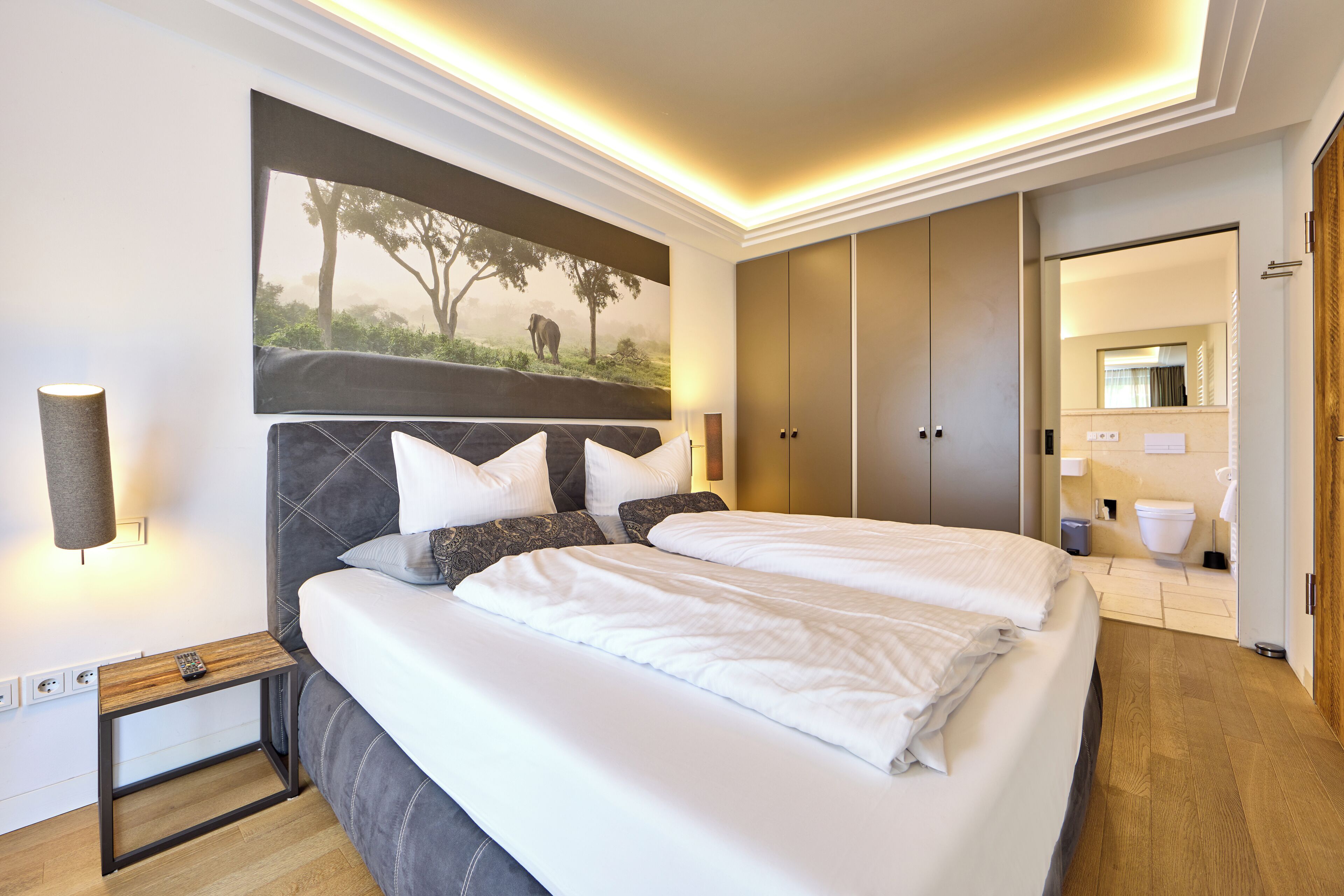 Ein modernes Hotelzimmer mit einem großen Bett, einem Kopfteil mit einer Naturmotiv-Abbildung, einem Nachttisch mit einer Lampe und einem Kleiderschrank.