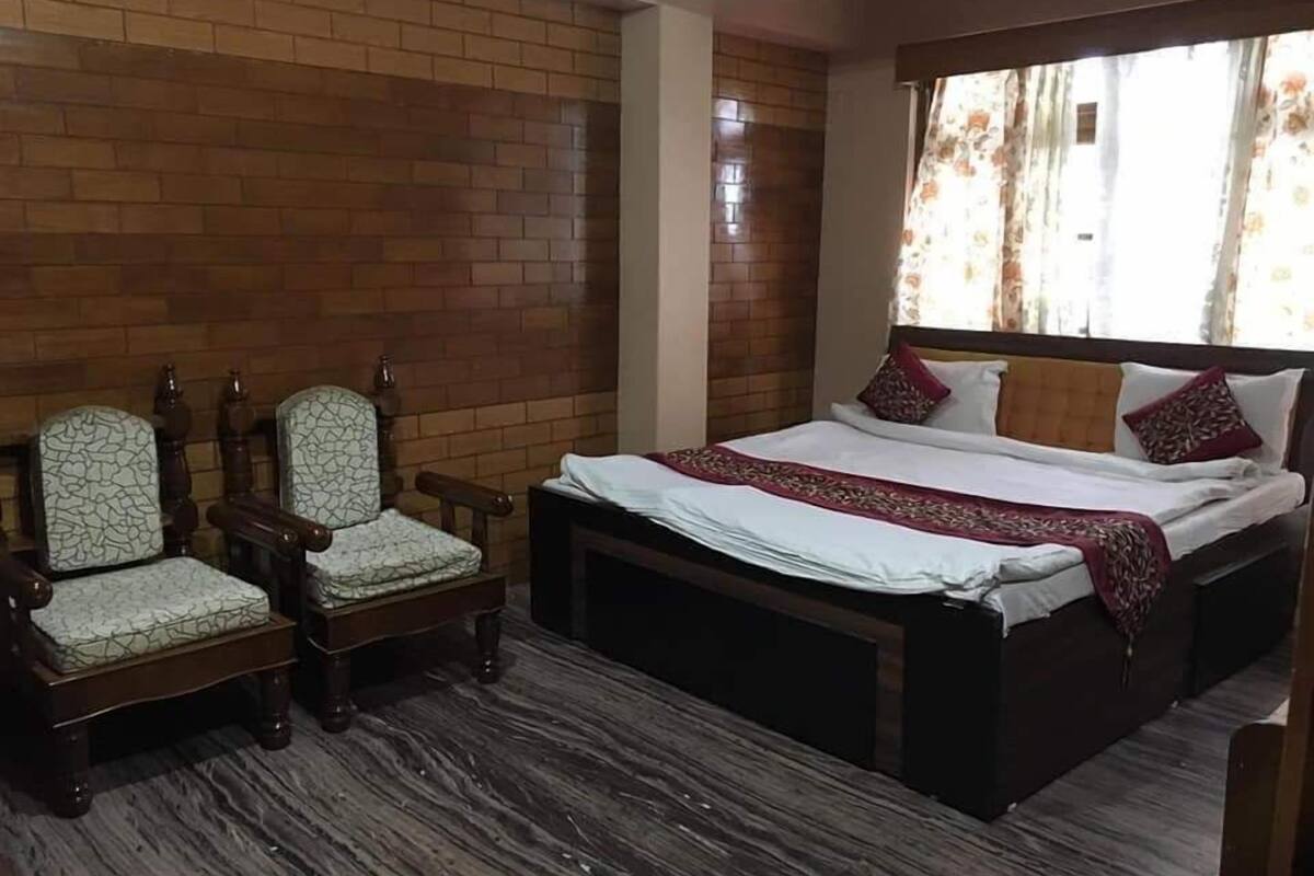 A bedroom with a brick wall, a bed with a white and red bedspread, two upholstered chairs, and a window with floral curtains.