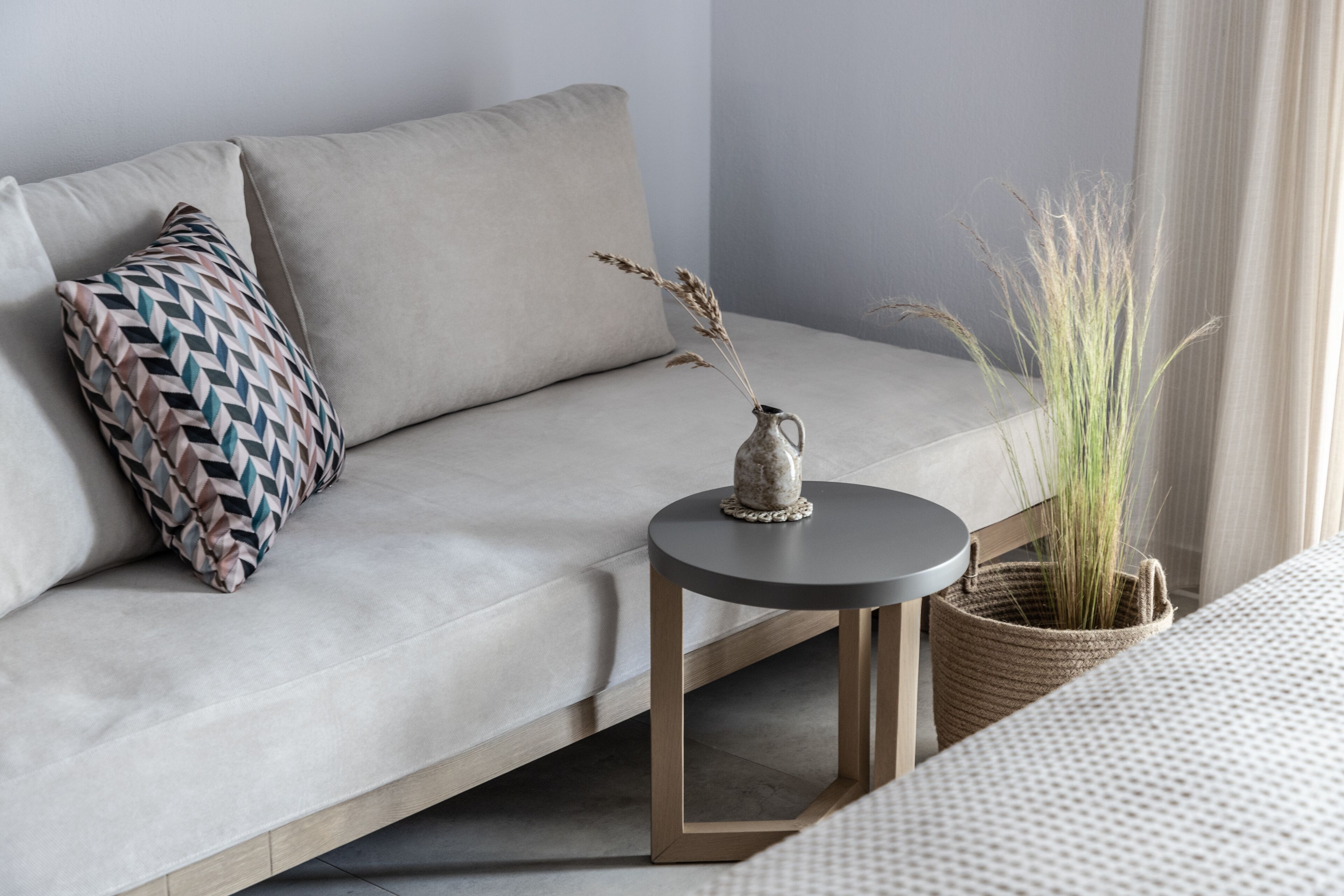 A modern living room with a sofa, a patterned pillow, a small round table with a vase of dried plants, and a woven basket with green plants.