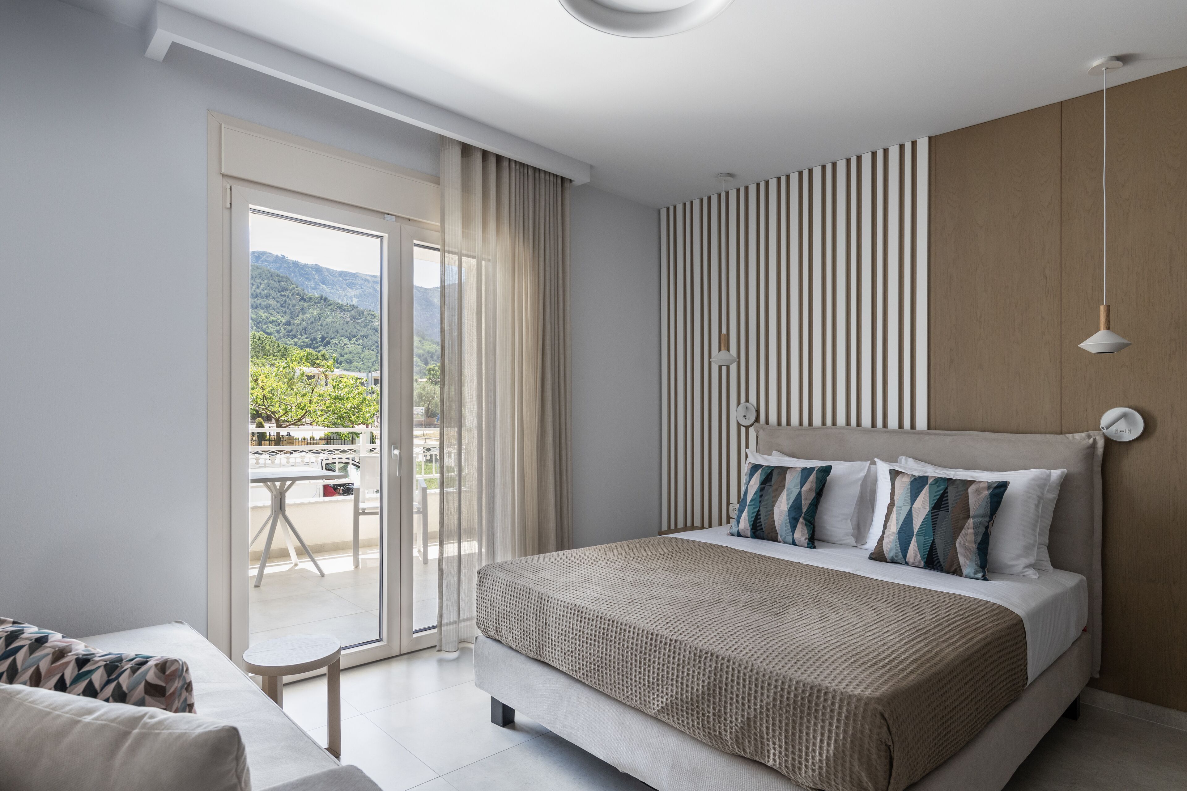 A modern bedroom with a large bed, a sliding glass door to a balcony, and a view of greenery.