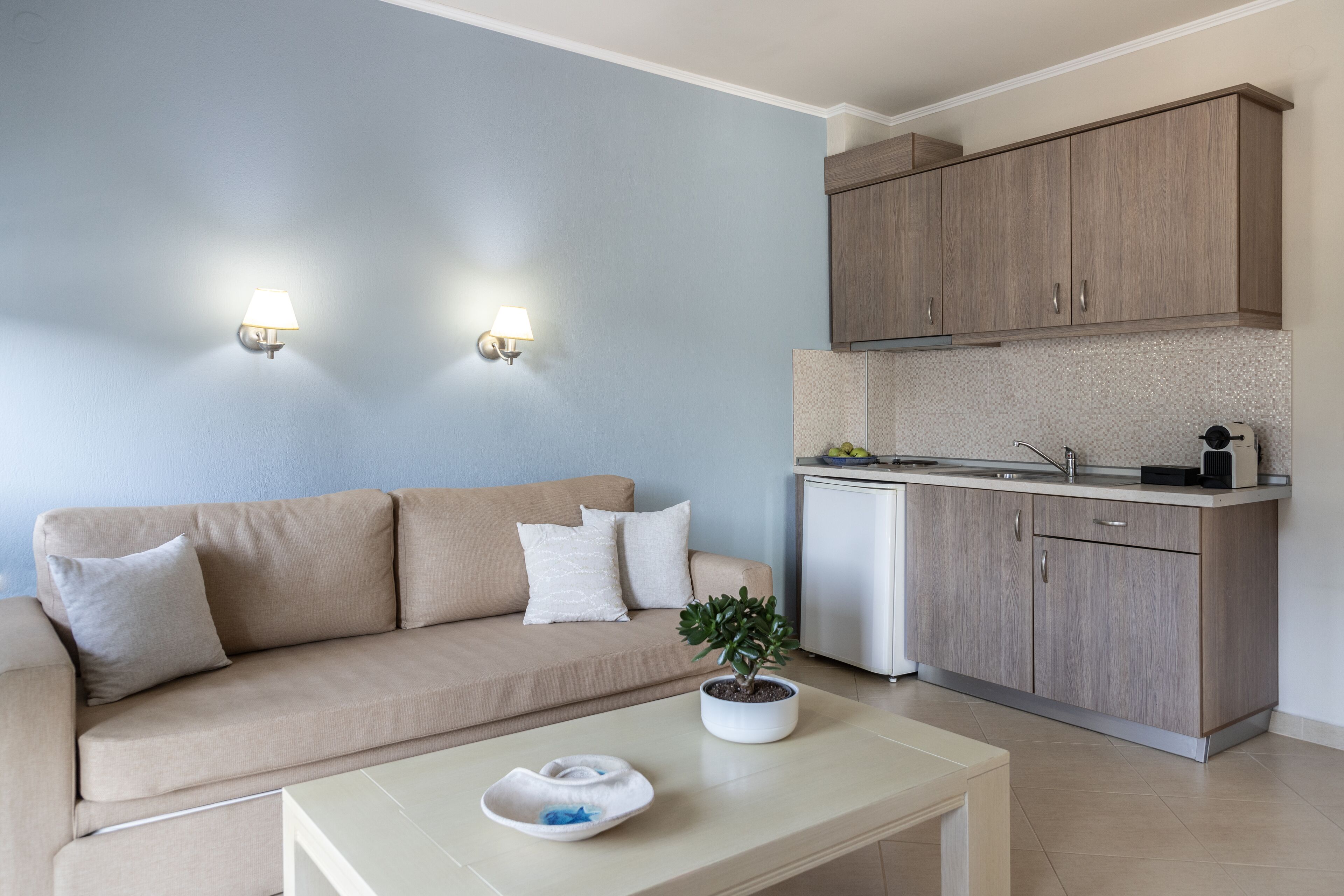 A modern living room with a beige sofa, two wall-mounted lights, a small potted plant, and a kitchen area with wooden cabinets and a white refrigerator.