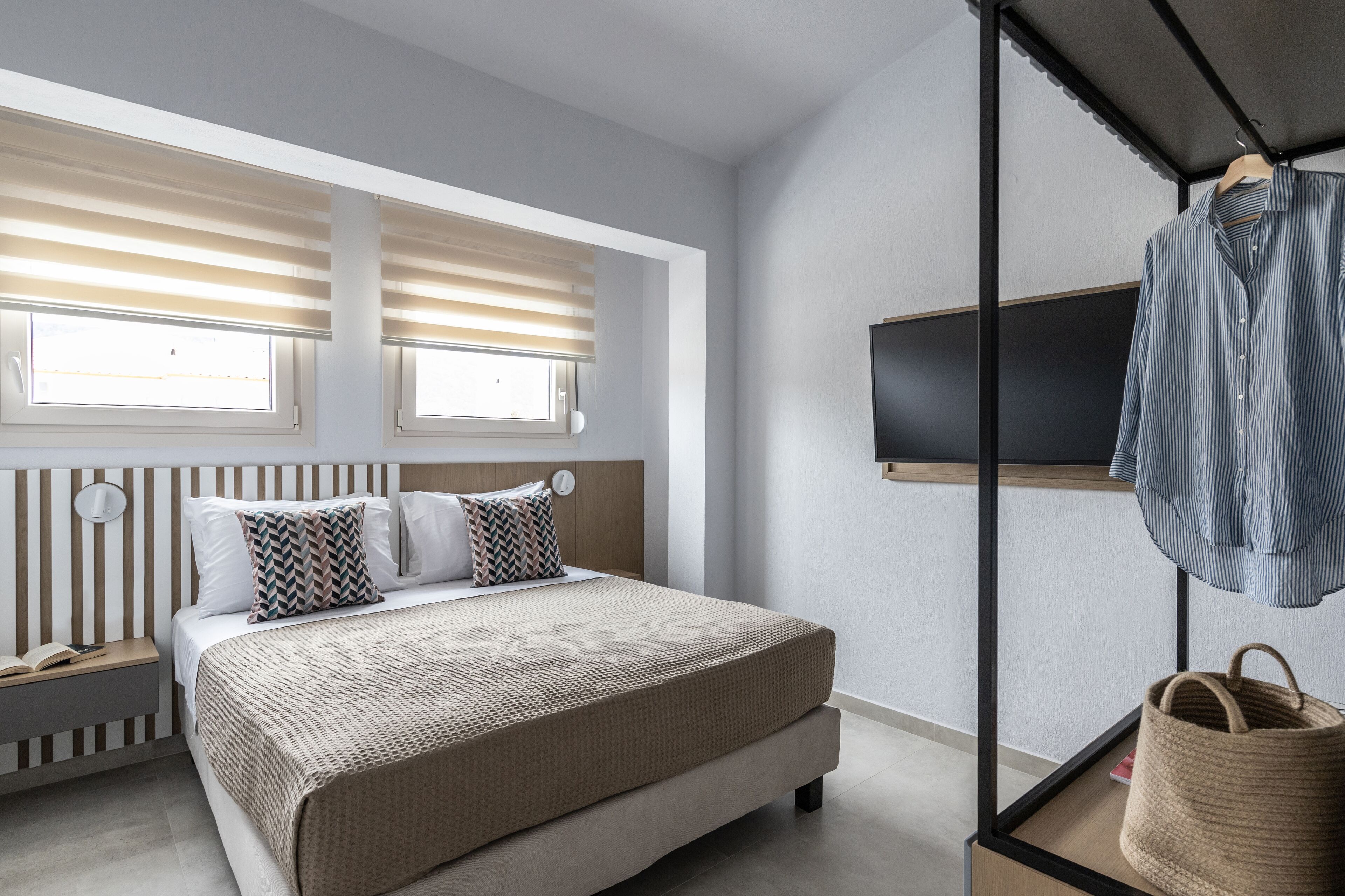 Un dormitorio moderno con cama, dos ventanas con persianas, una mesita de noche, televisor montado en la pared y un perchero.