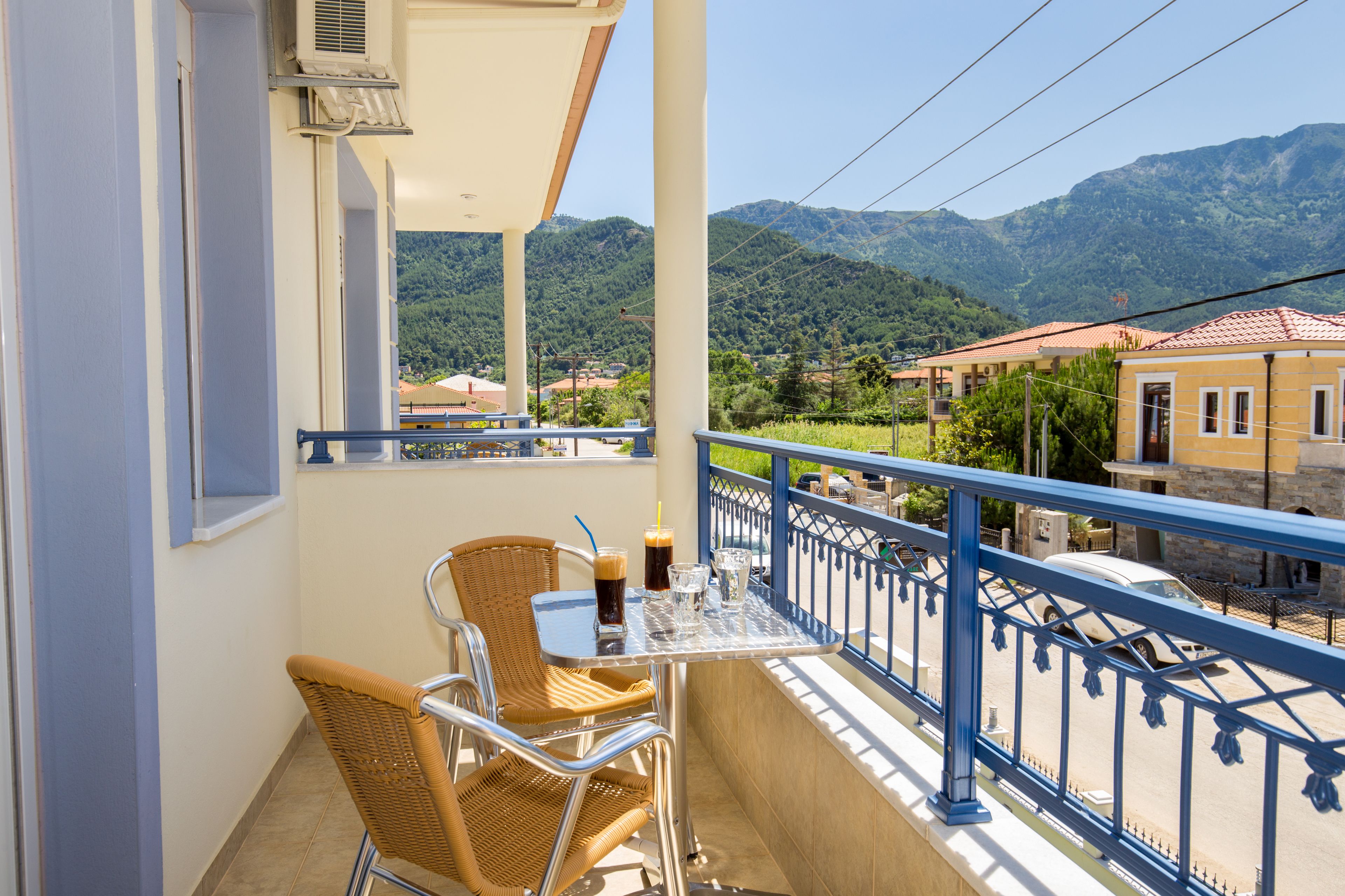 A balcony with wicker chairs, a table with drinks, and a view of mountains and buildings.
