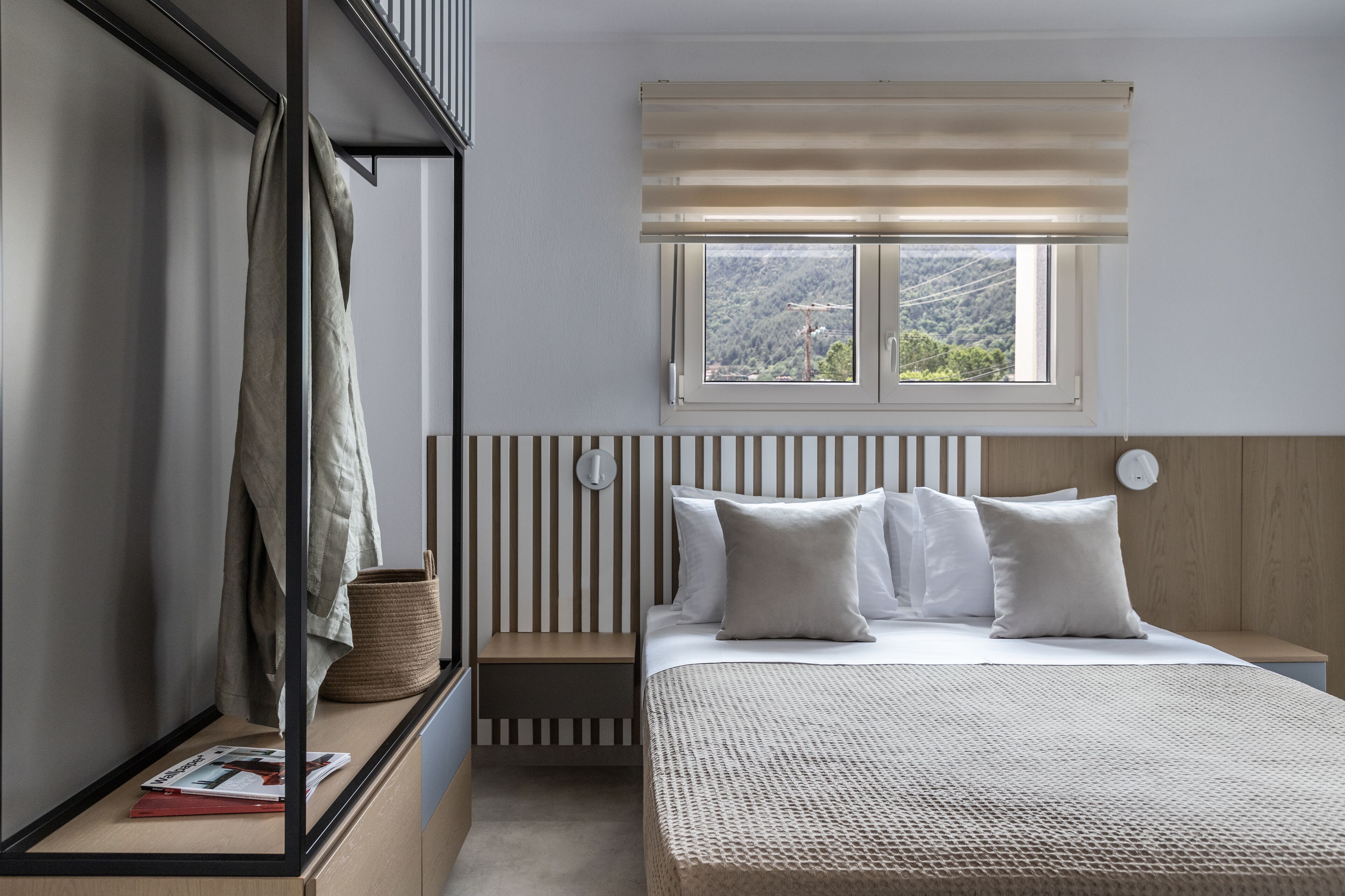 A bedroom with a bed, a nightstand, a window with blinds, a mirror, and a view of a forested area.