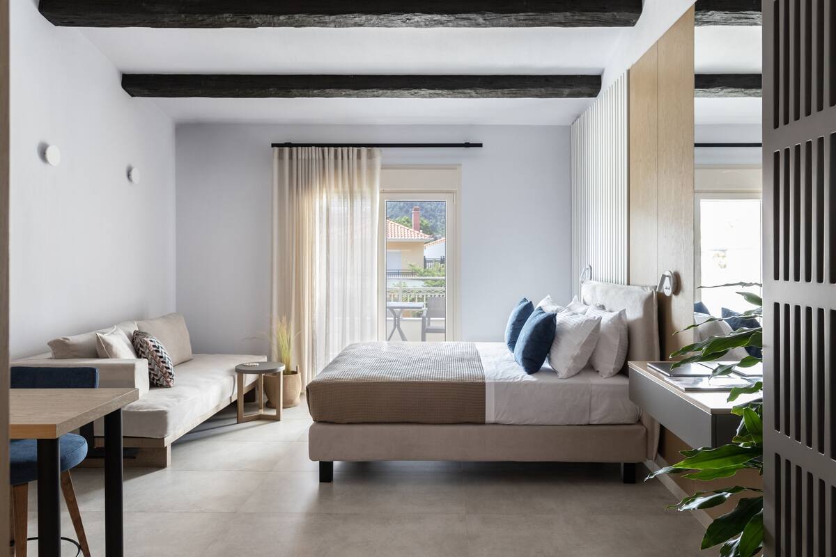 A modern hotel room with a bed, a sofa, a desk, and a view of the outdoors.