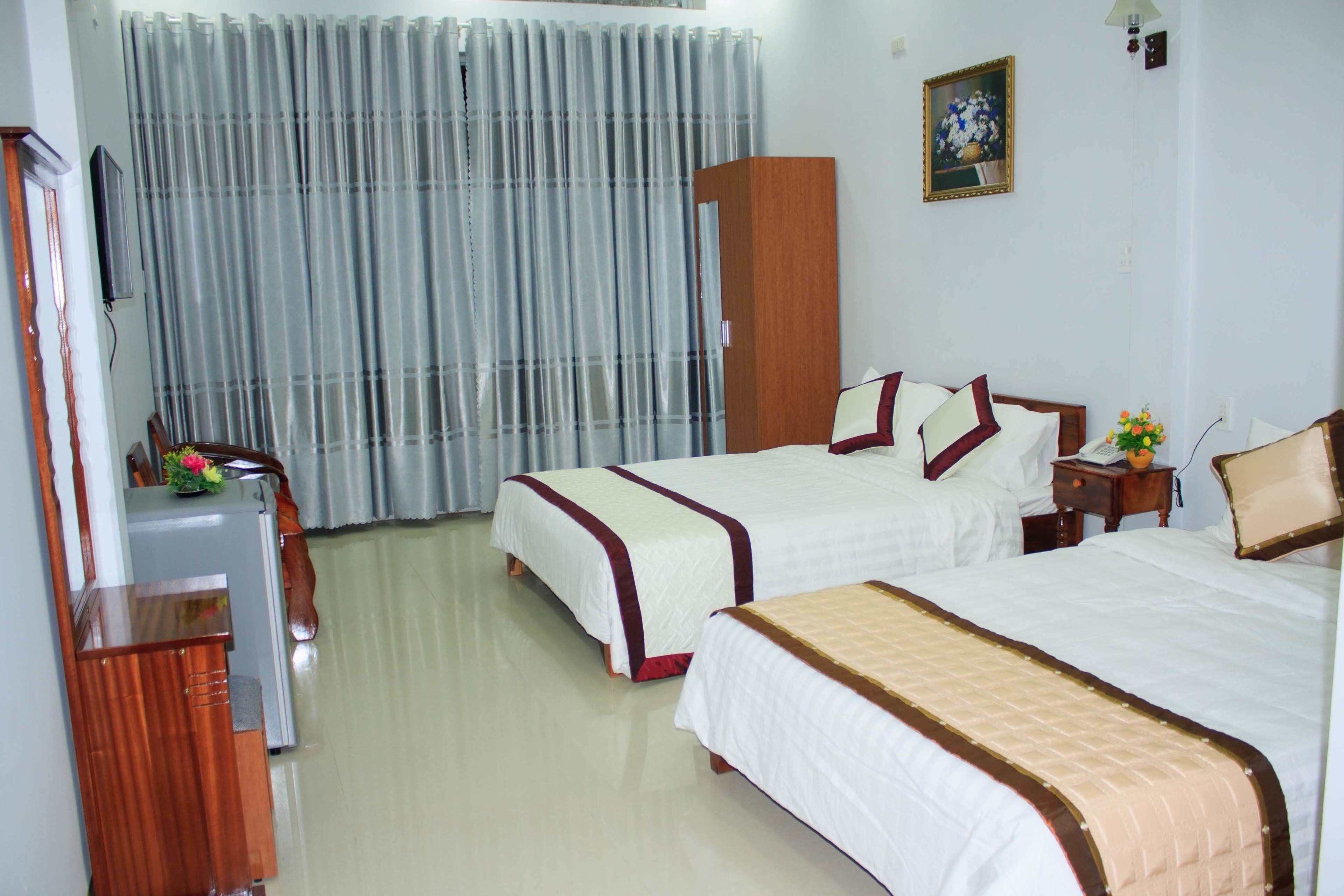 A hotel room with two single beds, a wooden desk, a small table with a vase, and a television on the wall.