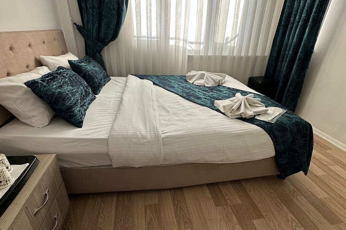 Una cama bien hecha con cabecera, un aparador de madera con cajones y una ventana con cortinas traslúcidas.