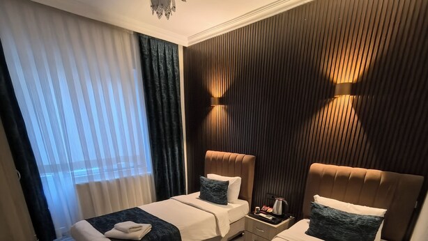 A hotel room with two beds, a ceiling fan, a nightstand with a lamp, and a view of the outside through sheer curtains. - Hotel Estambul (Istanbul)