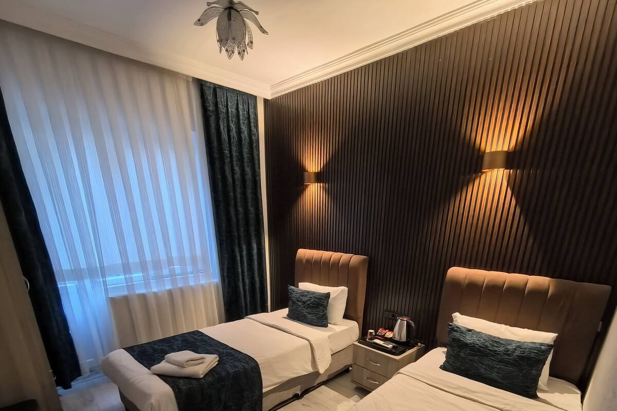 Habitación de hotel con dos camas, ventilador de techo, mesita de noche con lámpara y vistas al exterior a través de cortinas traslúcidas.