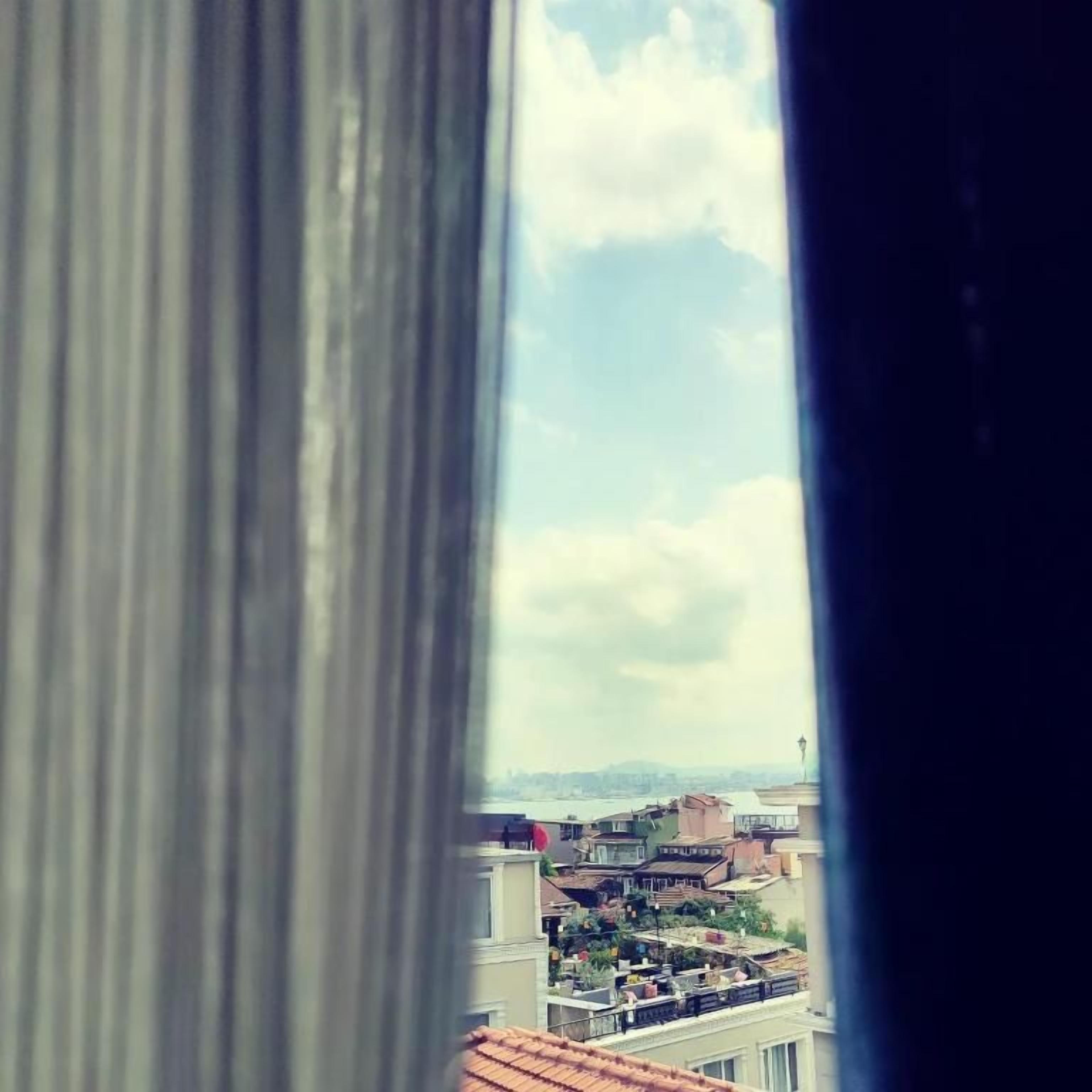 Blick durch ein Fenster mit Vorhang, der eine Stadtansicht mit Gebäuden und blauem Himmel zeigt.