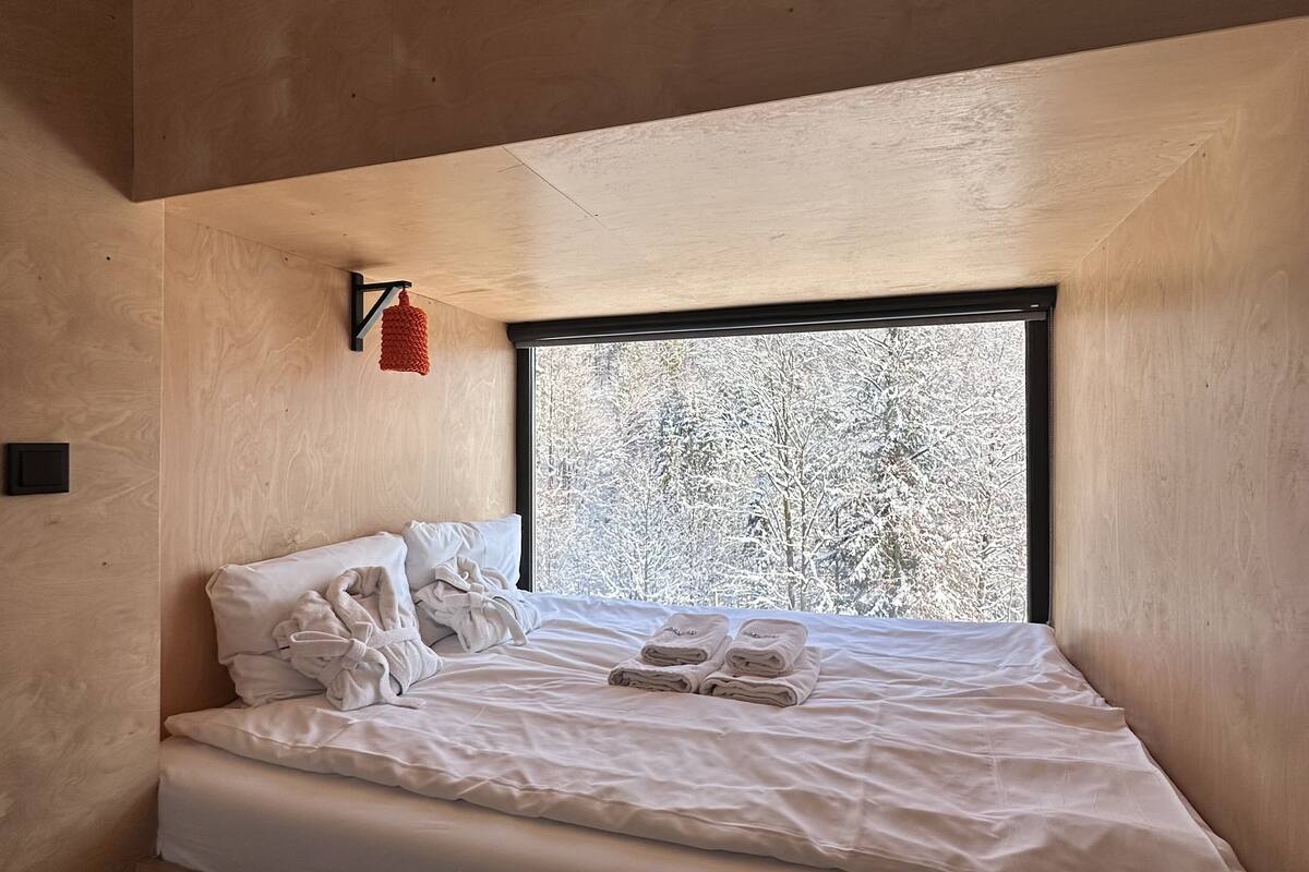 A small, cozy bedroom with a single bed, a window with a view of snow-covered trees, and a red hanging light fixture.