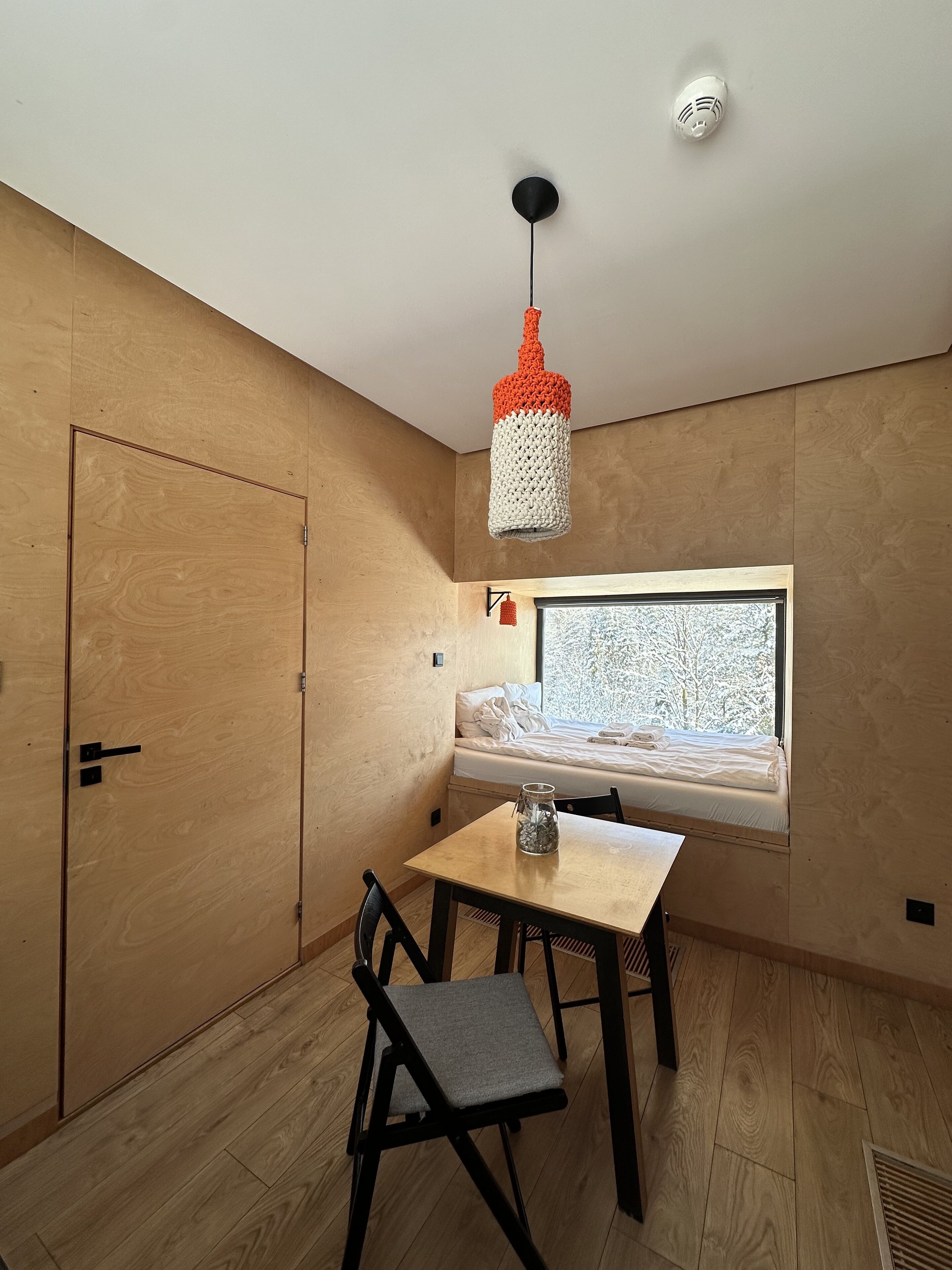 A room with a wooden door, a window seat with a view, a table with a chair, and a hanging light fixture.