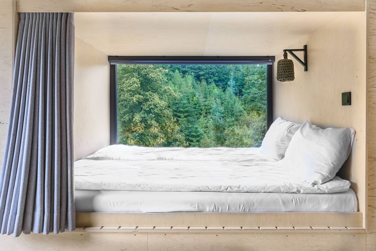 A bed with a view of a forest through a large window.