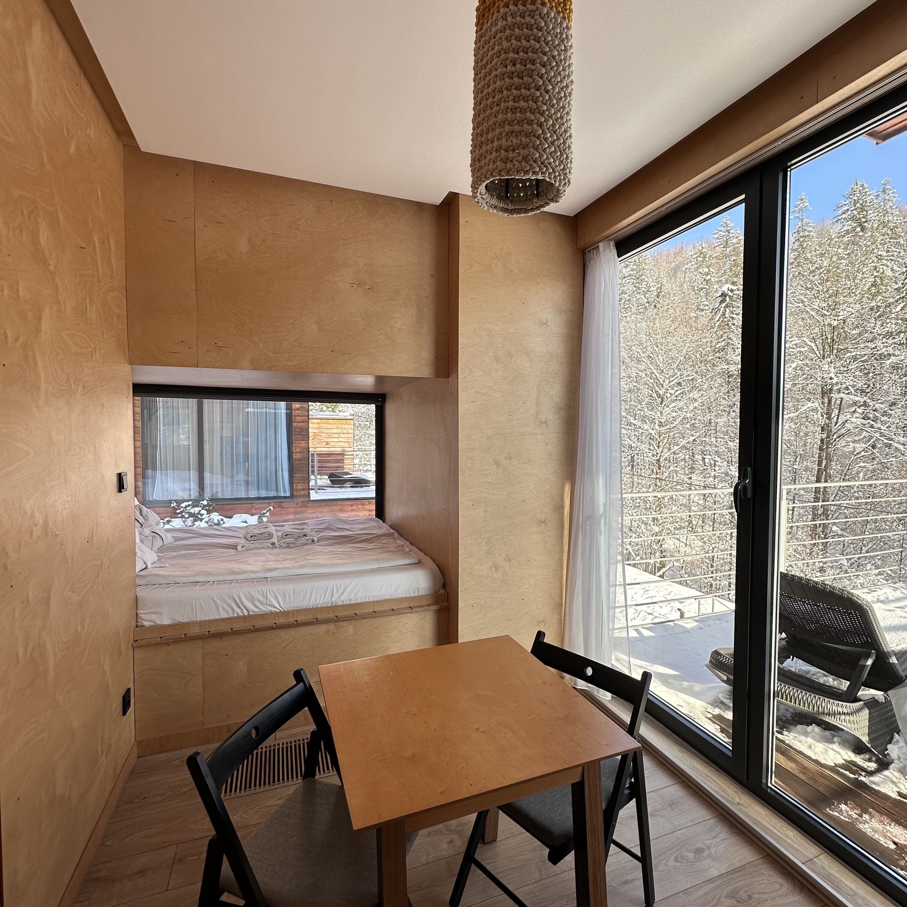 Un dormitorio pequeño con cama, una mesa y sillas, y un ventanal con vistas a árboles cubiertos de nieve.