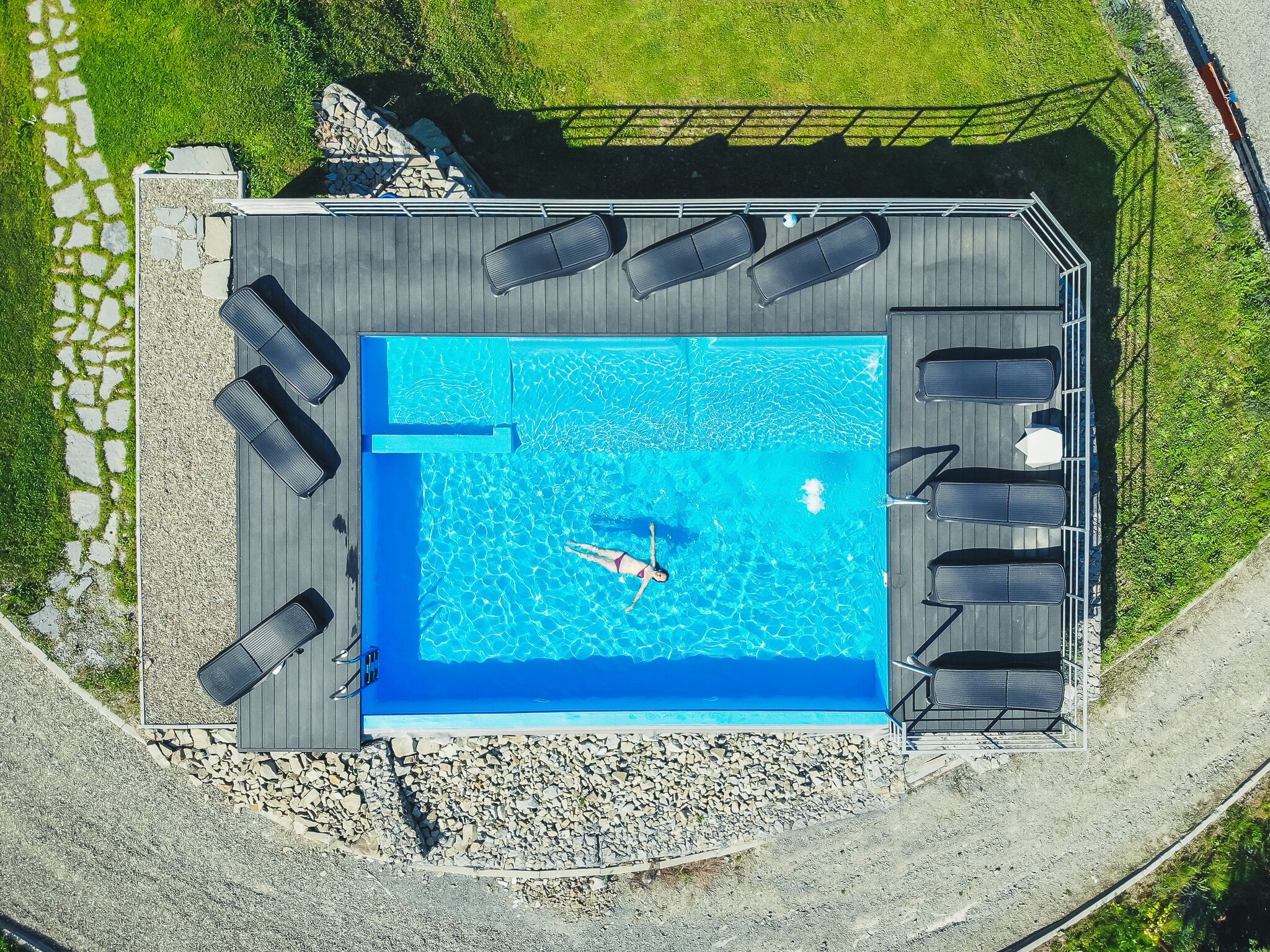 An aerial view of a rectangular swimming pool with a deck, surrounded by a stone wall and a grassy area.