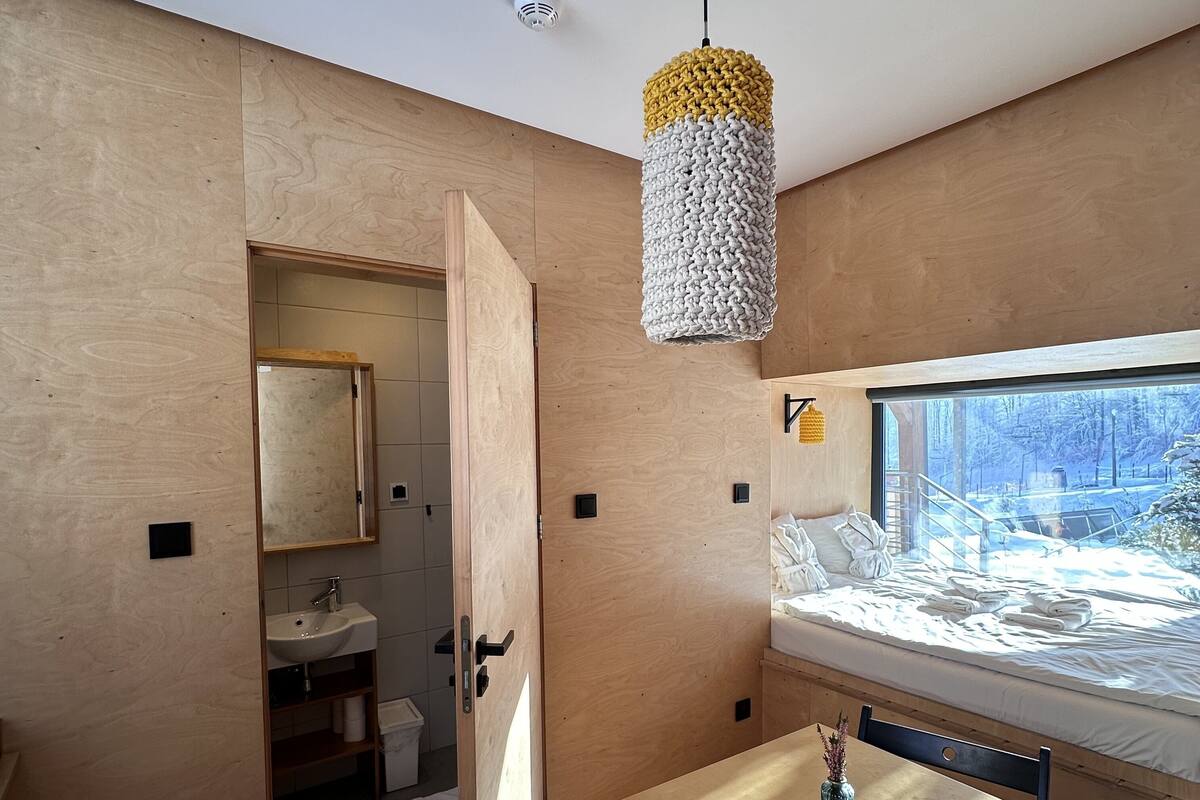 A modern room with a wooden interior, a bed with a view of a snowy landscape, a small table with a vase, and a hanging light fixture.