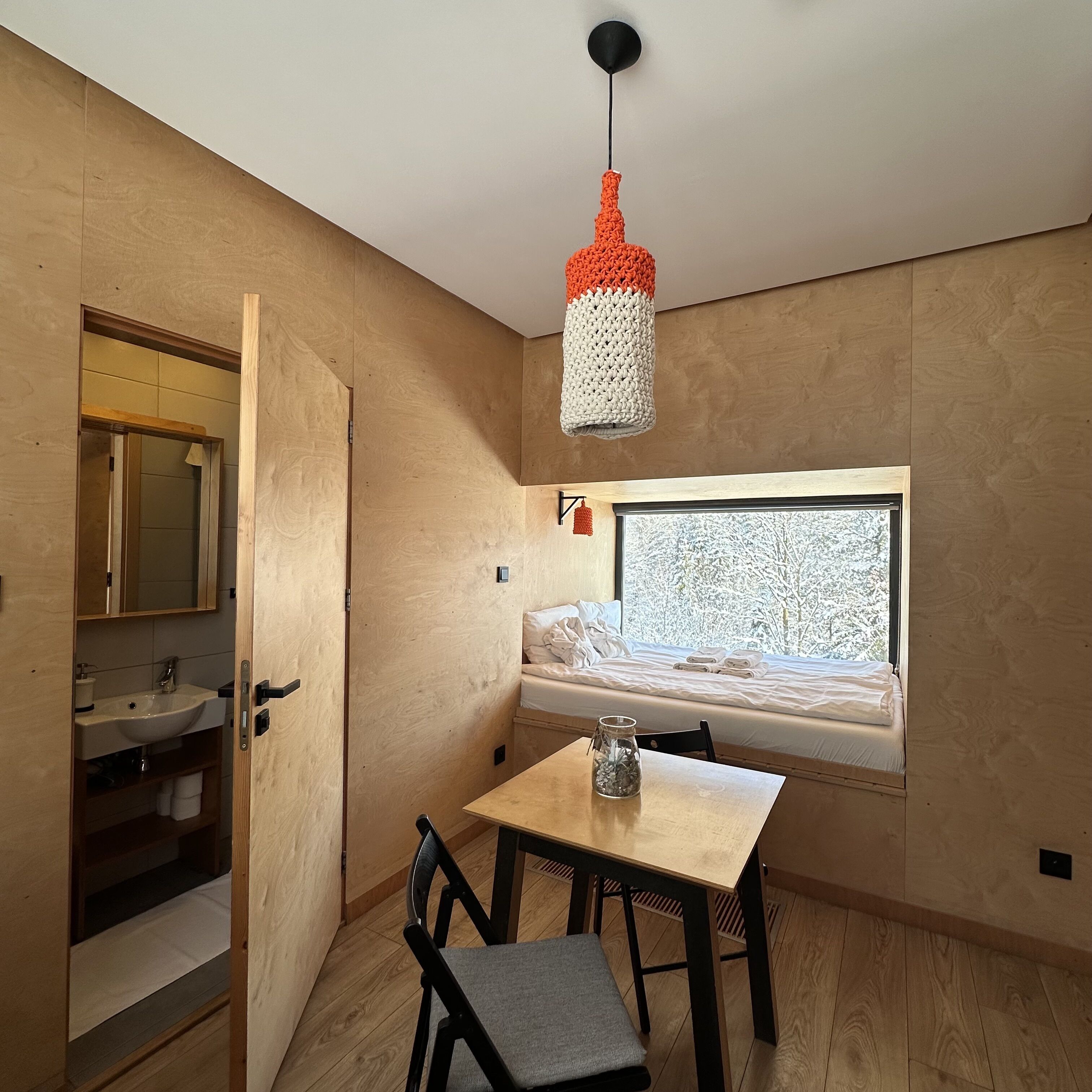A modern room with a wooden floor, a small table with a chair, a bed with a view of snow-covered trees, and a hanging light fixture.