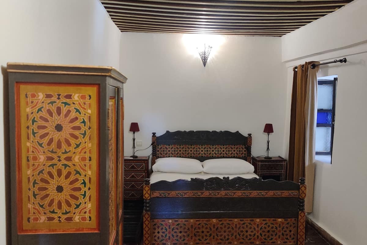 A bedroom with a bed, a nightstand, a lamp, a wardrobe, and a patterned rug.