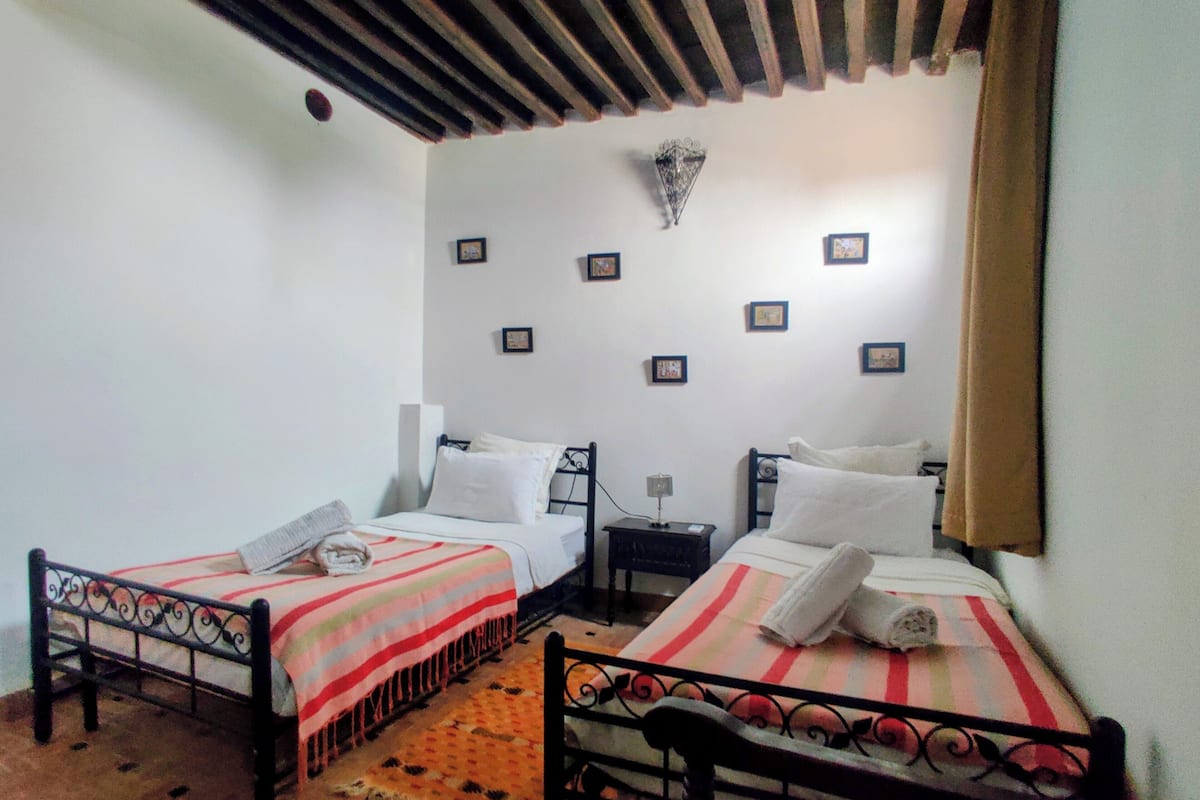 A room with two beds, each with a striped bedspread and pillows, a small table with a lamp, and a wall-mounted light fixture.