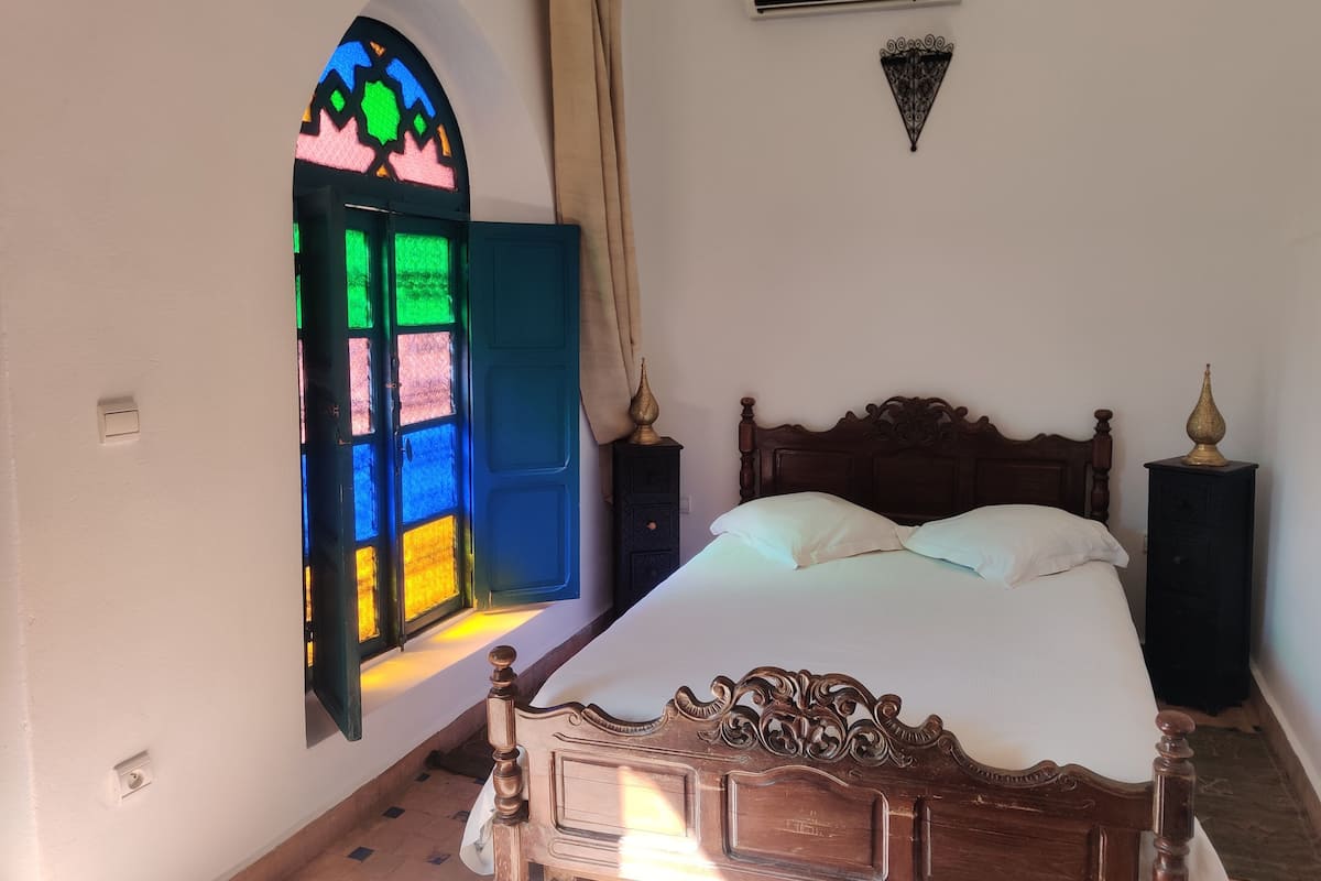A bedroom with a wooden bed, a stained glass window, an air conditioning unit, and a decorative wall piece.