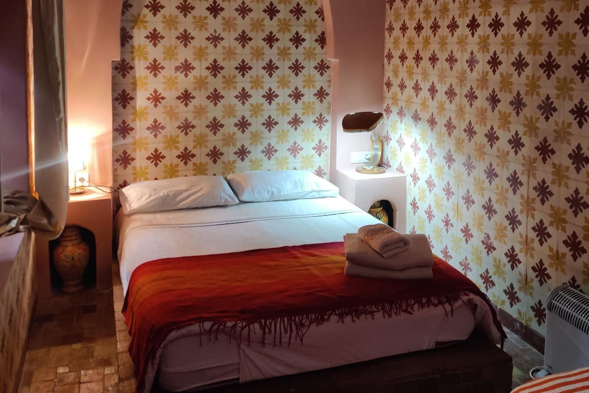 A room with a bed, a nightstand, a lamp, a patterned wall, and a painting.
