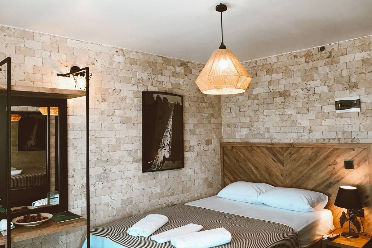 A hotel room with a brick wall, a wooden headboard, a bed with white linens, a hanging light fixture, and a mirror.
