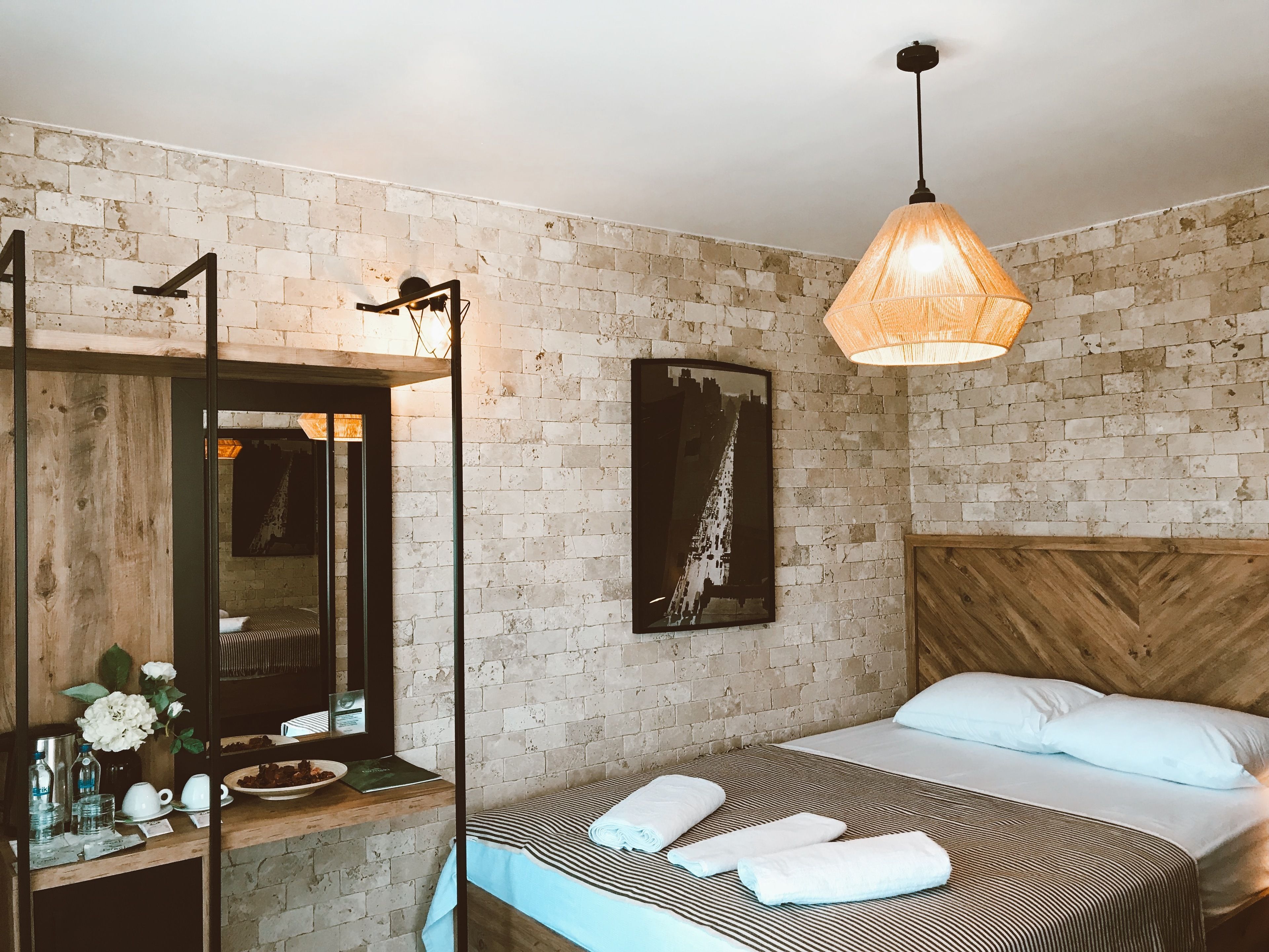 Een slaapkamer met een bakstenen muur, een bed met witte kussens, een houten hoofdbord, een hangende lamp en een spiegel met houten lijst.
