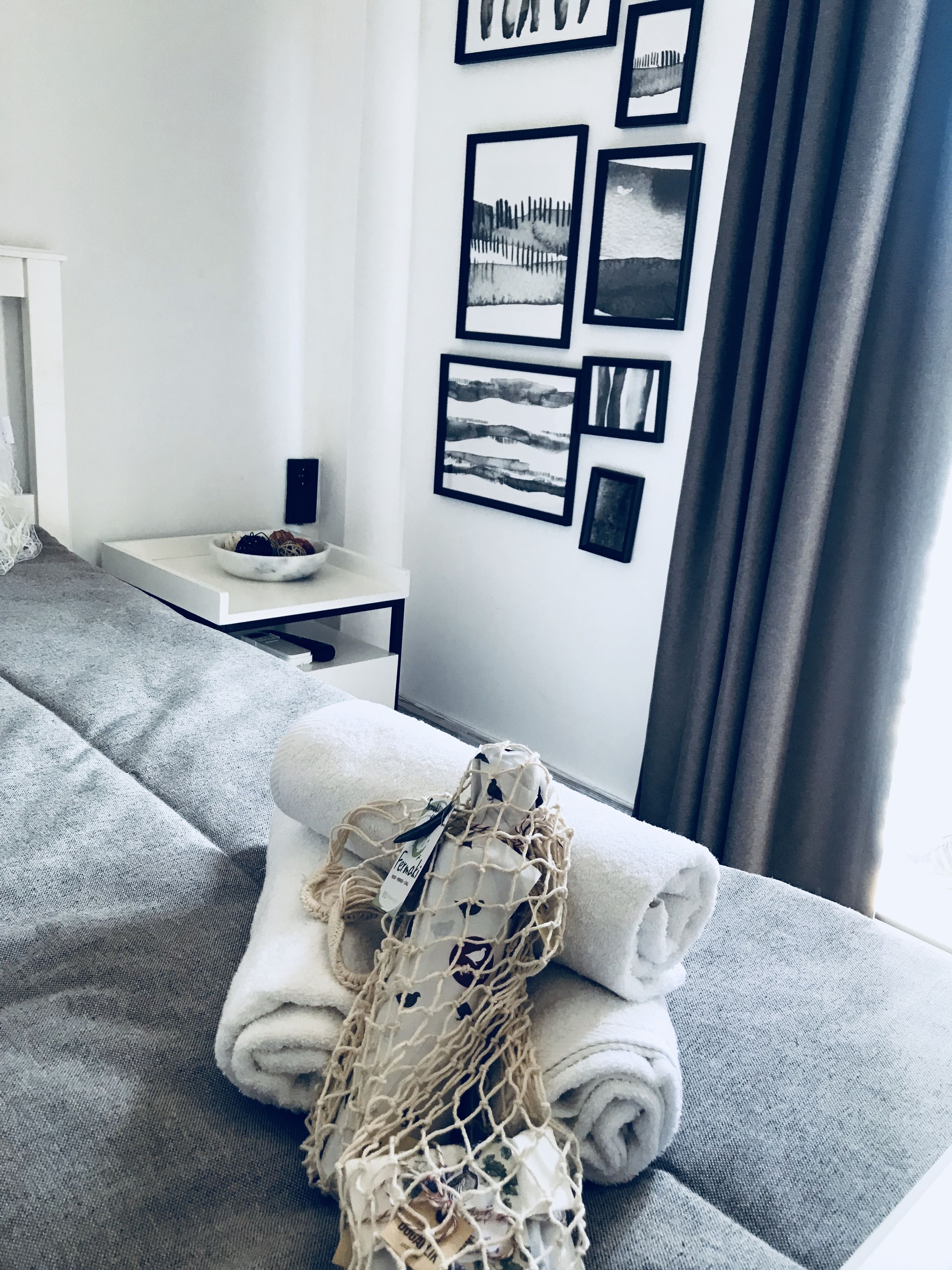 A neatly made bed with a grey blanket, a white towel, and a netted bag on a grey sofa. There are framed pictures on the wall and a small table with a bowl and a bottle.