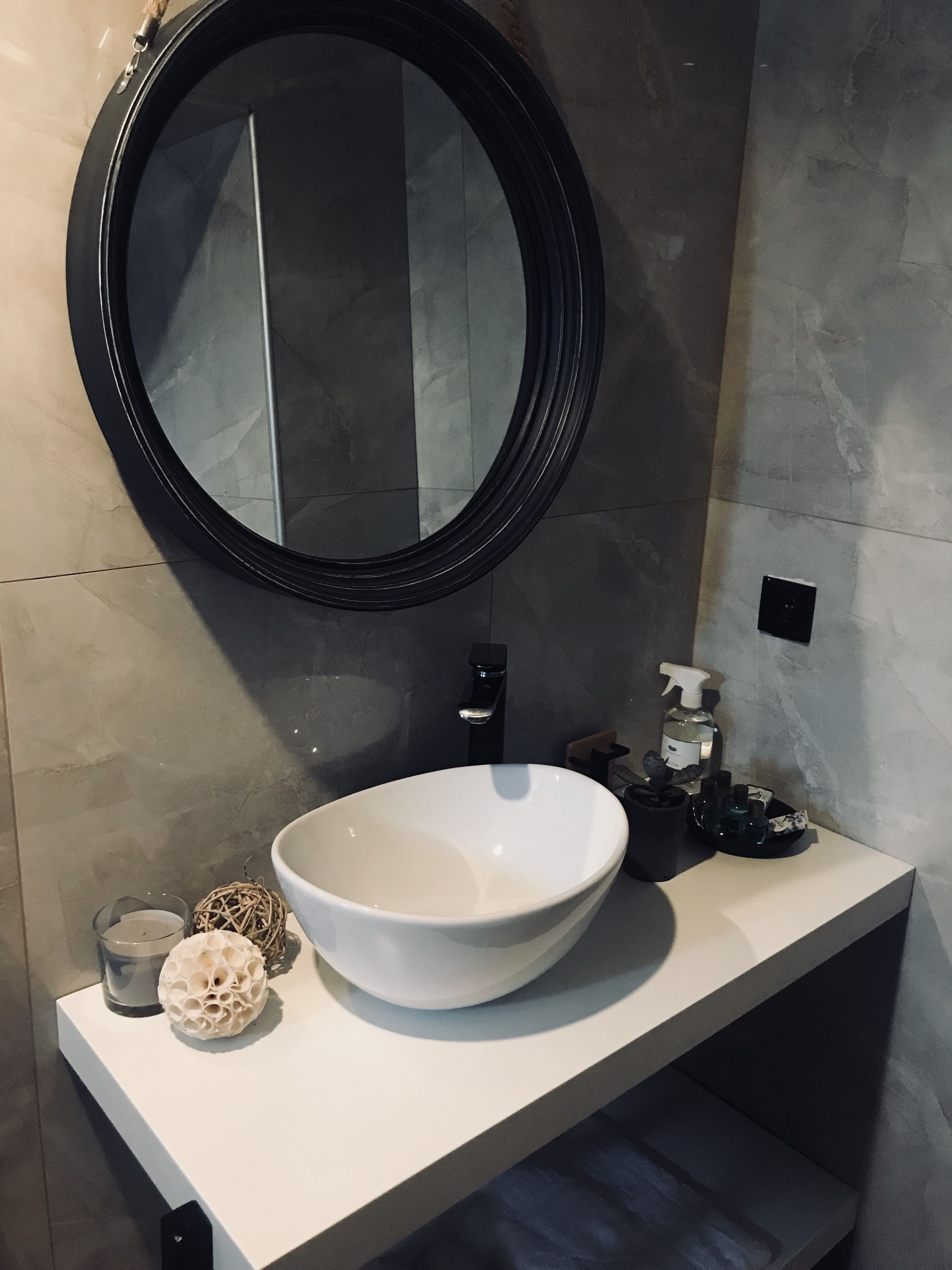 A bathroom with a round mirror, a white sink, a black faucet, a soap dispenser, and a tissue holder.