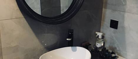 Un baño con un espejo redondo, un lavabo blanco, un grifo negro, un dispensador de jabón y un porta pañuelos.