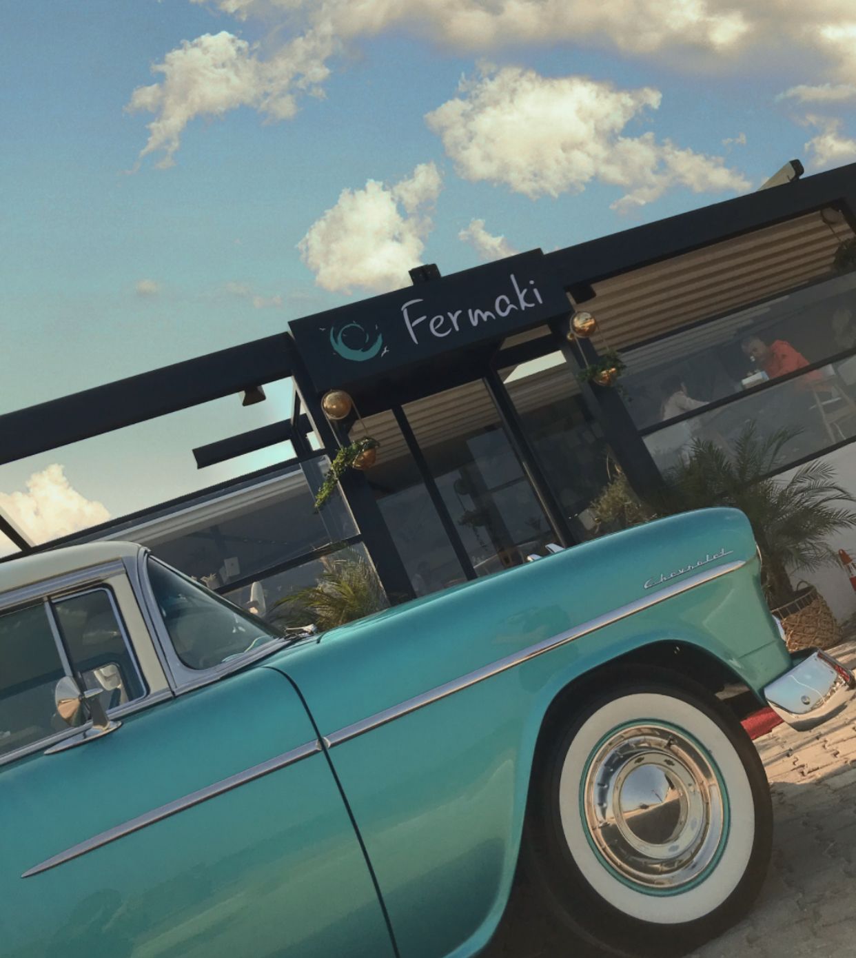 A teal vintage car parked in front of a building with a sign that reads "Fermaki."