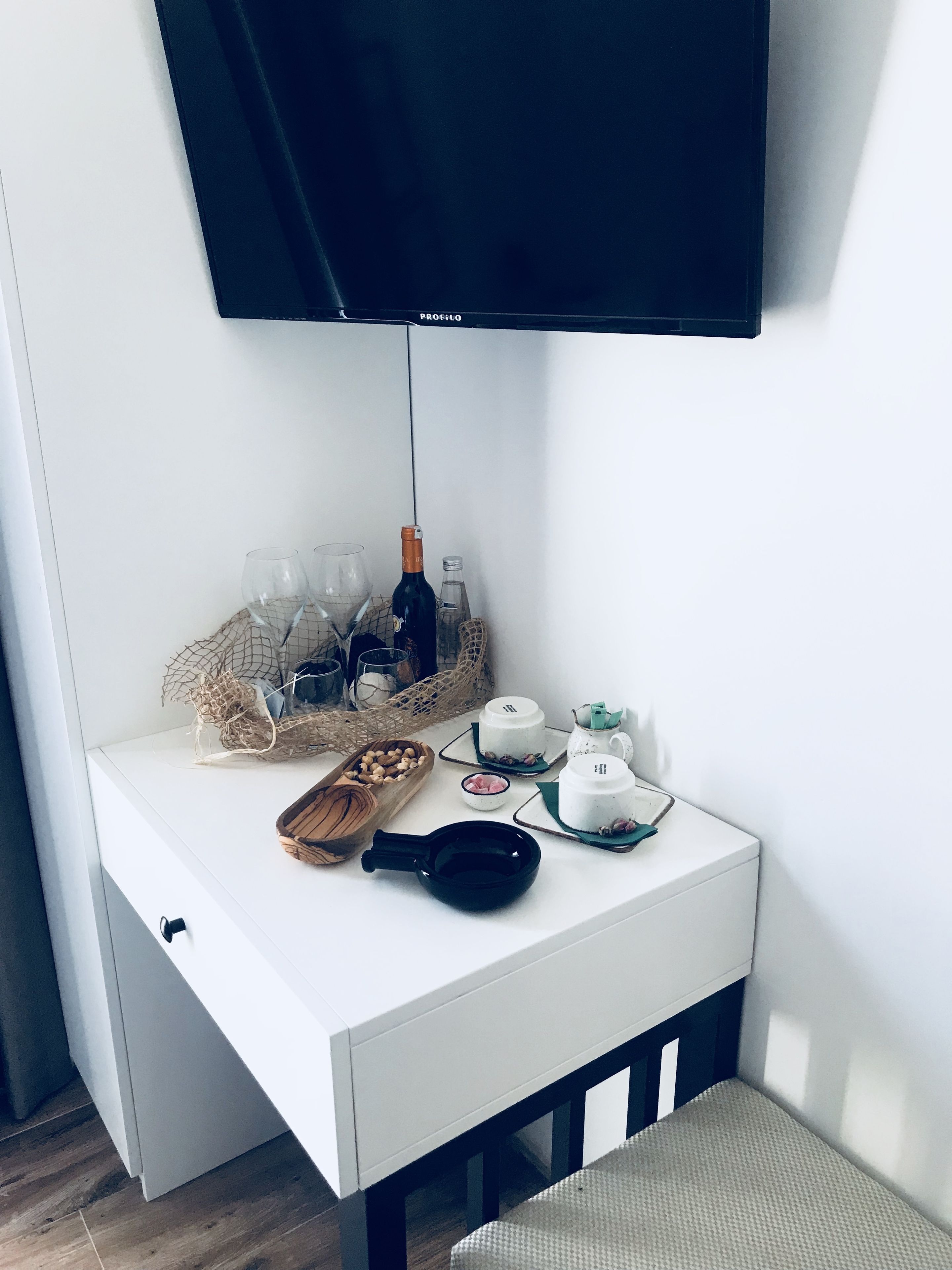Et hvidt natbord med et fladskærms-tv monteret over. Bordet har en flaske, glas, en tekedel, en kop, en skål med nødder og en sort stegepande.