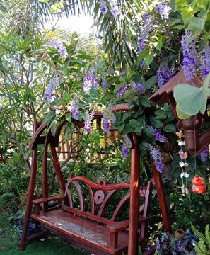 A wooden swing set with a bench, surrounded by lush greenery and purple flowers. - Tippawan Resort (Pattaya)