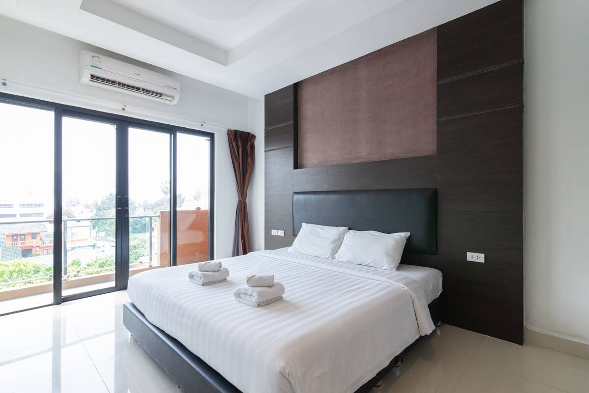Een moderne hotelkamer met een groot bed, een donker hoofdeinde en uitzicht op de stad door grote ramen.