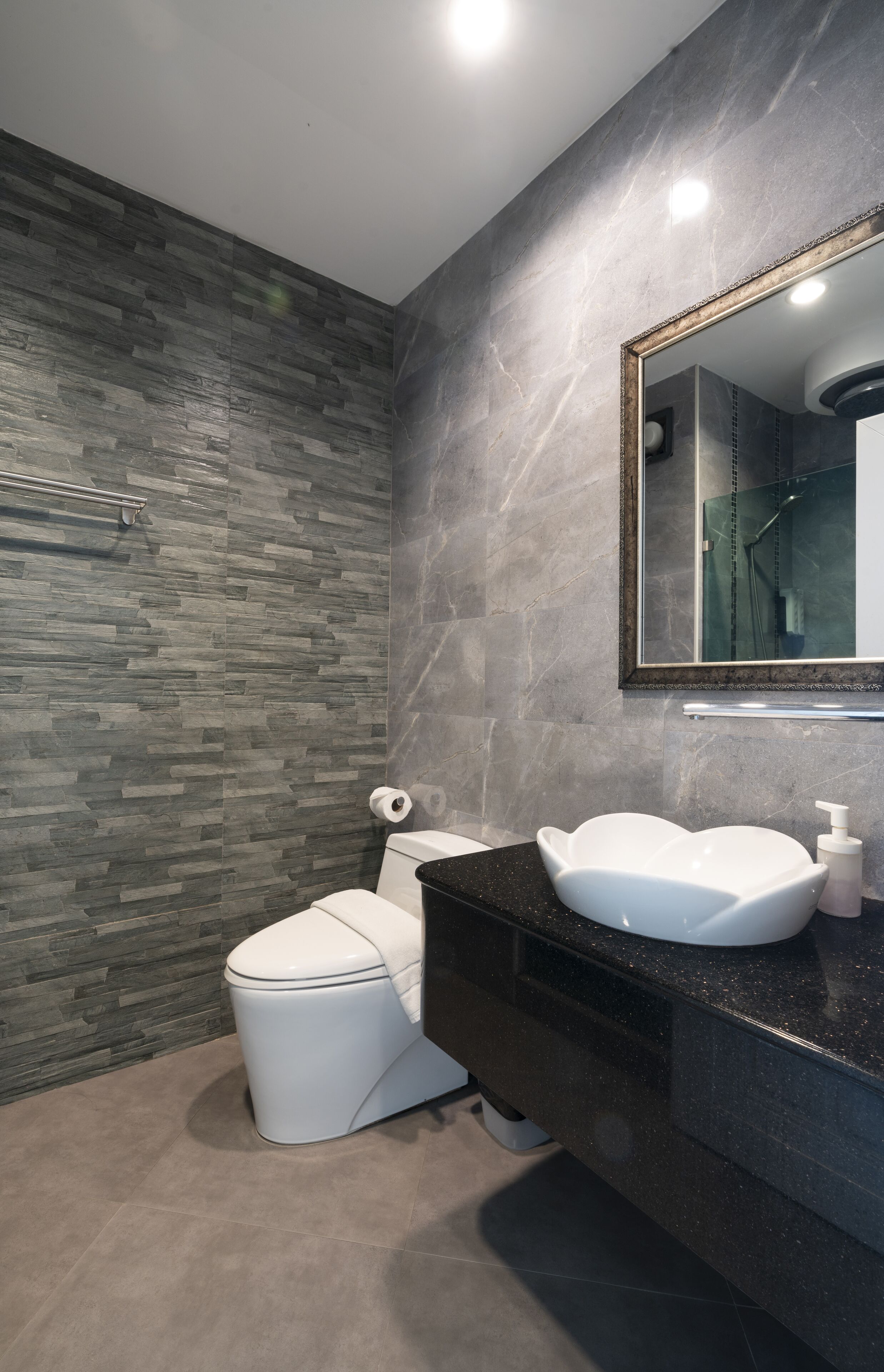 A modern bathroom with a stone wall, a white toilet, a black vanity with a white sink, and a mirror with a gold frame.