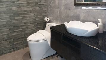A modern bathroom with a stone wall, a white toilet, a black vanity with a white sink, and a mirror with a gold frame.