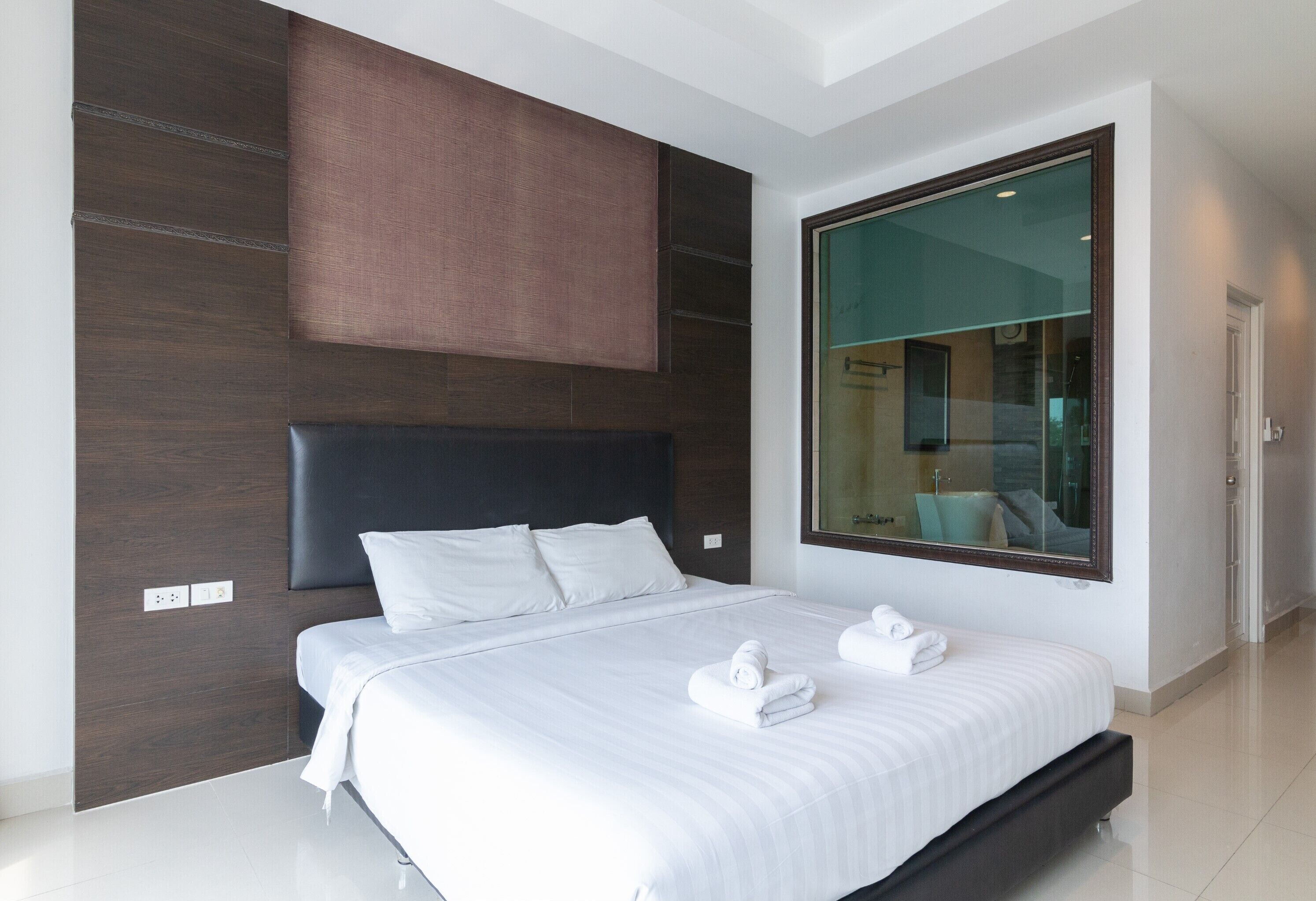 Ein modernes Hotelzimmer mit einem großen Bett, dunklen Holzpaneelen, einem großen Fenster mit Blick auf das Badezimmer und einem großen Spiegel.