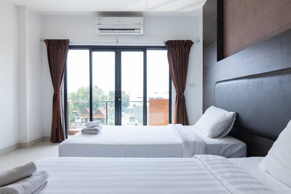 Een hotelkamer met twee bedden, een groot raam met gordijnen, een airconditioning en uitzicht op gebouwen.