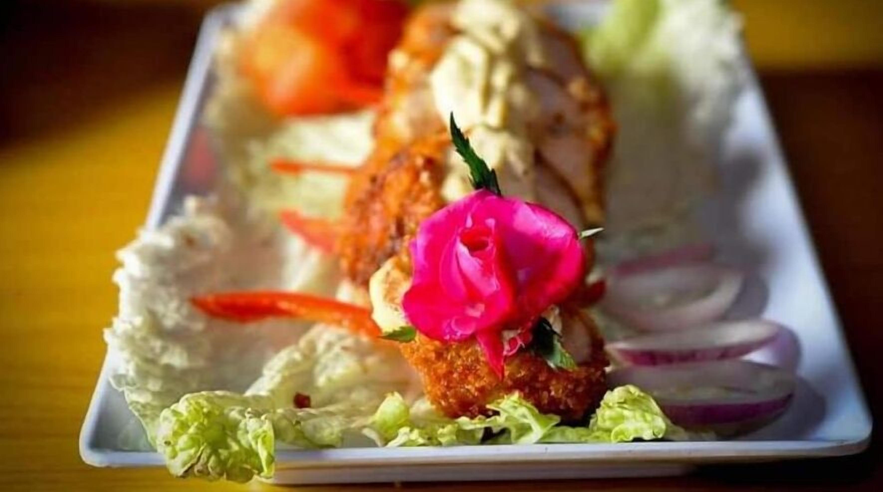 Un plato con un filete empanizado, posiblemente de pollo, adornado con una flor rosada y rodajas de cebolla roja, acompañado de lechuga y salsa cremosa.