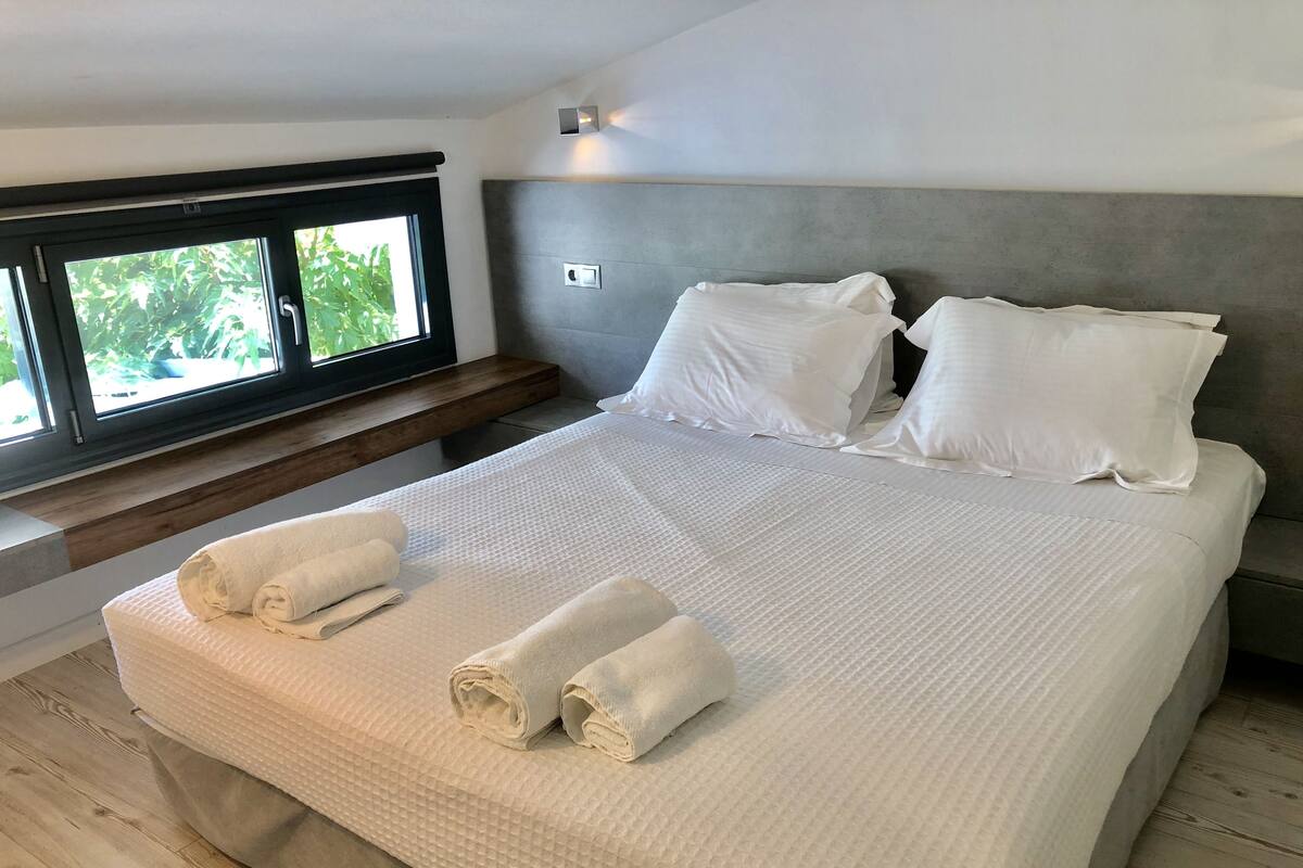A neatly made bed with white linens and towels, a wooden headboard, and a window with a view of greenery.