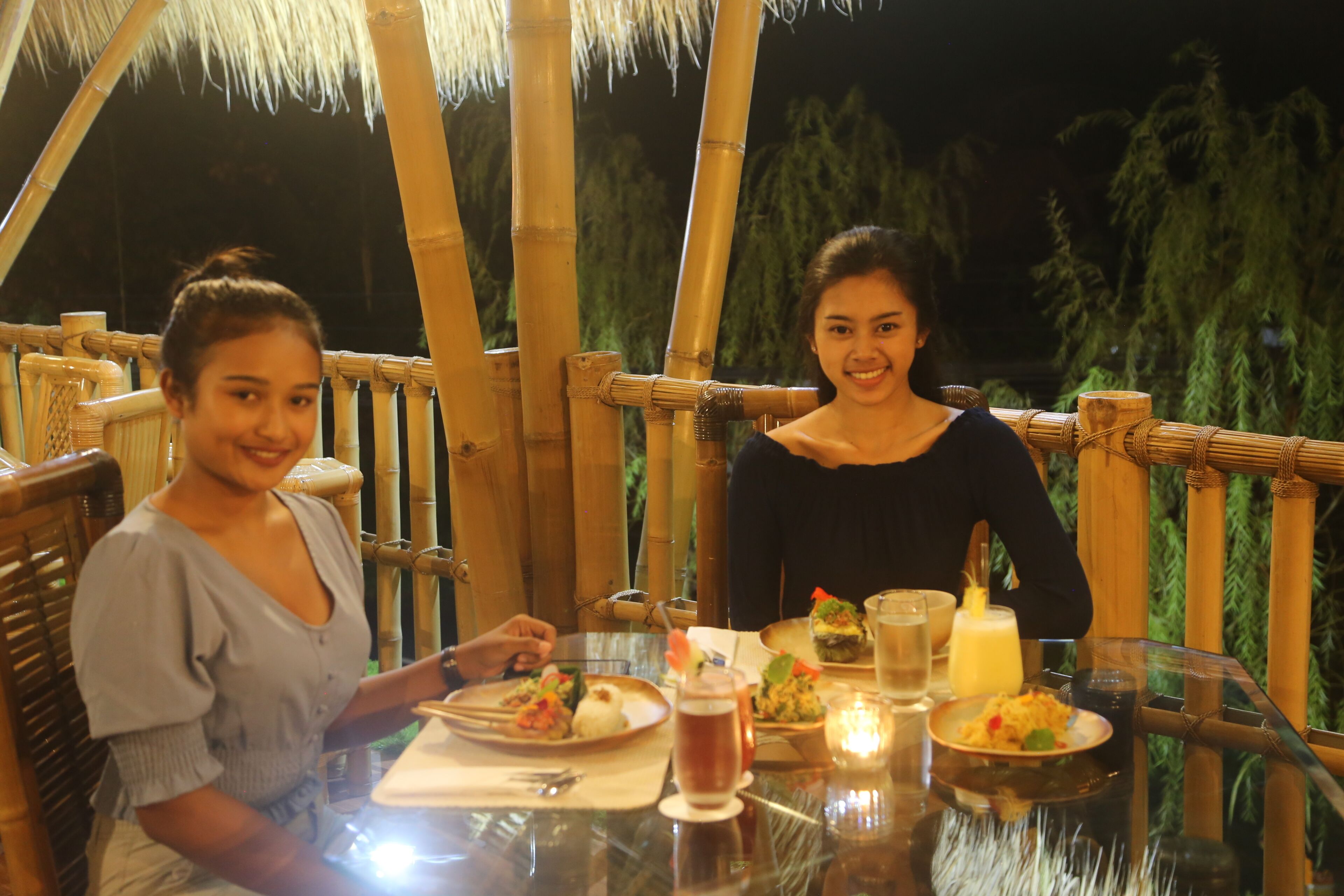Dos personas cenando en una mesa al aire libre, con estructuras de bambú y follaje de fondo.