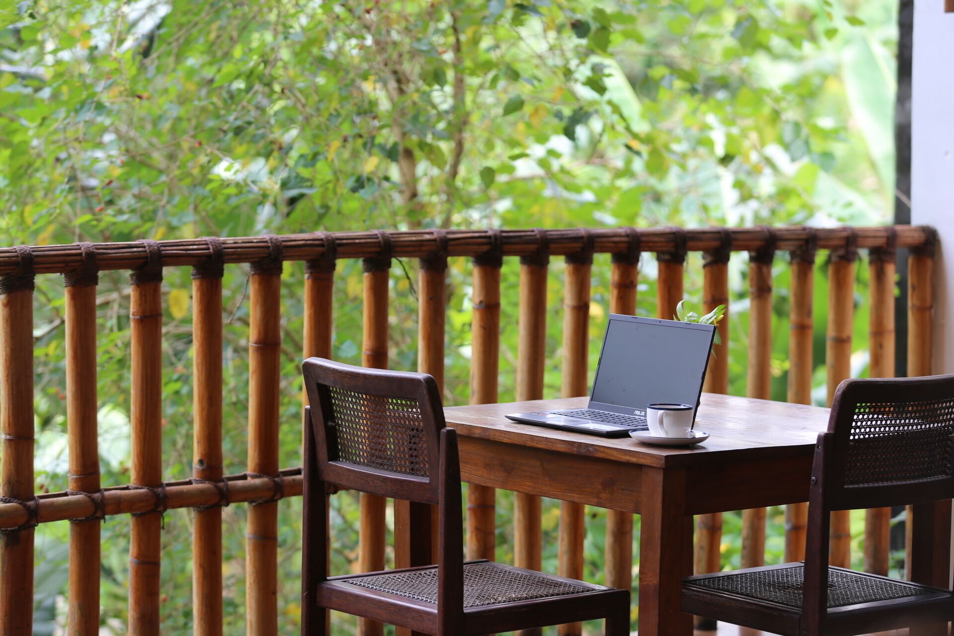 Une table en bois avec un ordinateur portable et une tasse, deux chaises en osier et une rambarde en bambou.