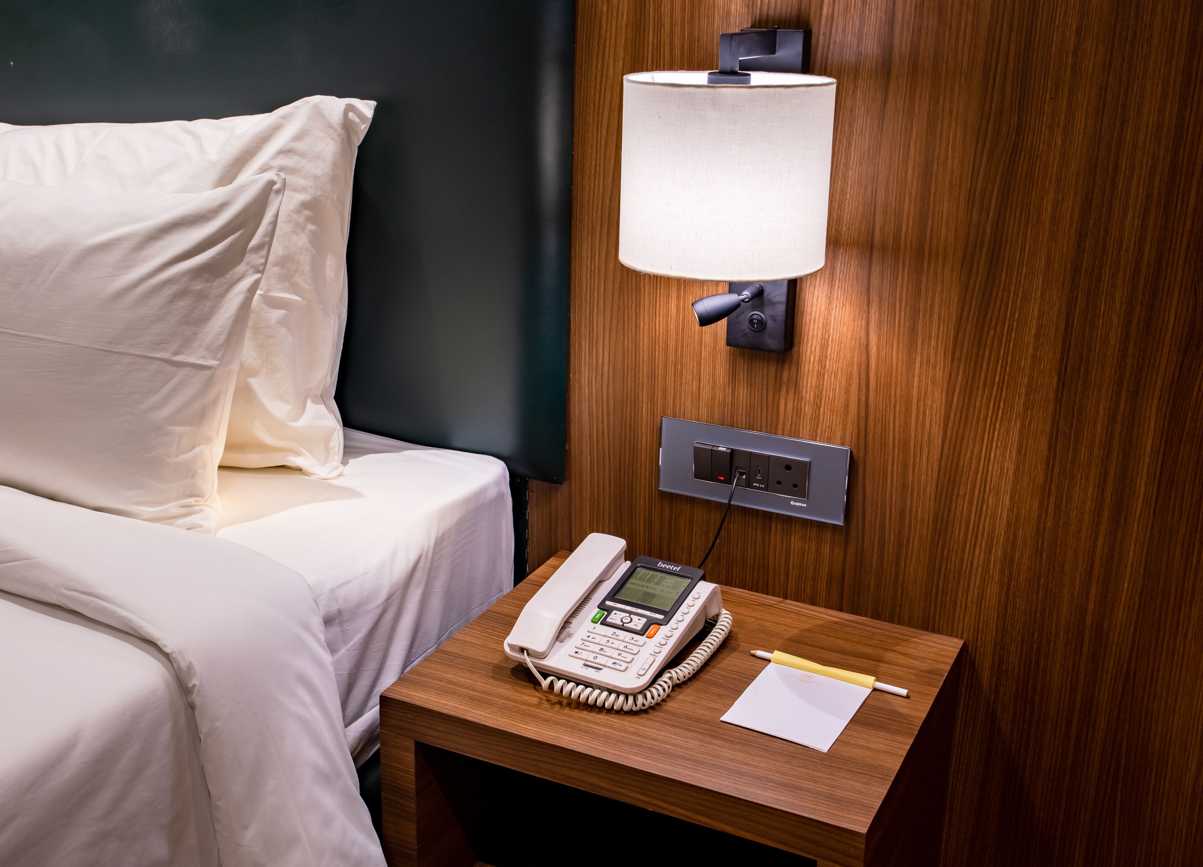 A hotel room with a bed, a bedside table with a phone, a lamp, and a power outlet.