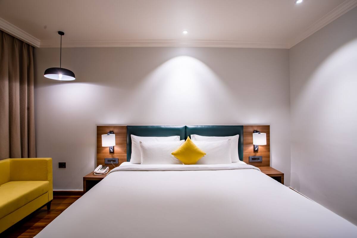 Une chambre d’hôtel avec un grand lit, un fauteuil jaune, deux lampes de chevet et un luminaire au plafond.