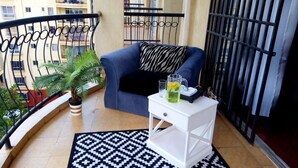 Un balcon avec un canapé bleu, une petite table de chevet blanche, un tapis à motifs, une plante en pot et une rambarde en métal.