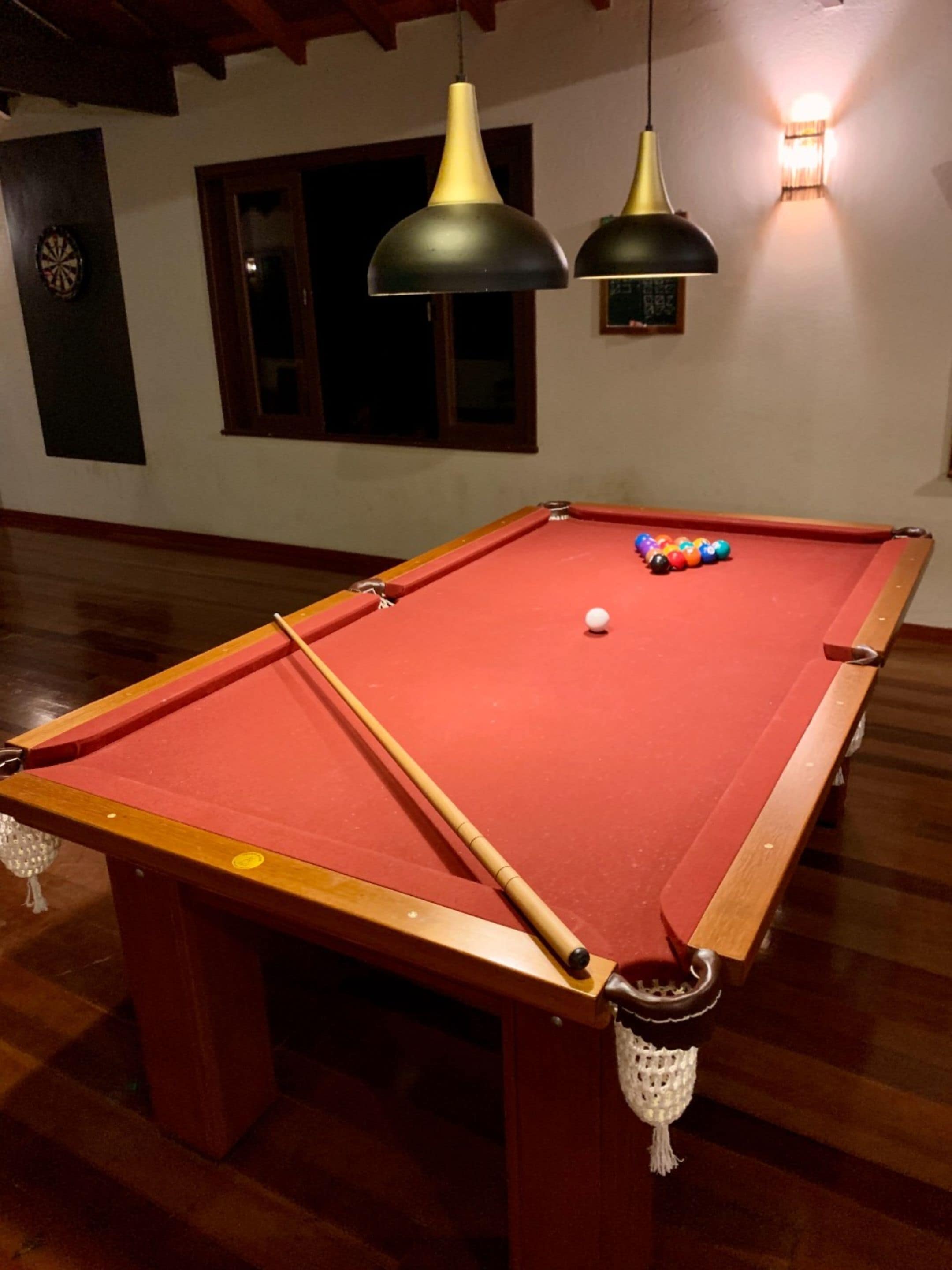 Een biljart met een rode viltkleed, een keu en kleurrijke ballen. Boven de tafel hangen twee hanglampen.