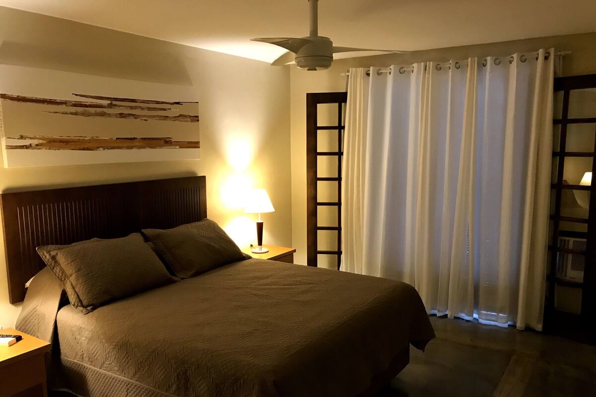 A neatly made bed with a headboard, a bedside lamp, a nightstand, a ceiling fan, and a window with curtains.