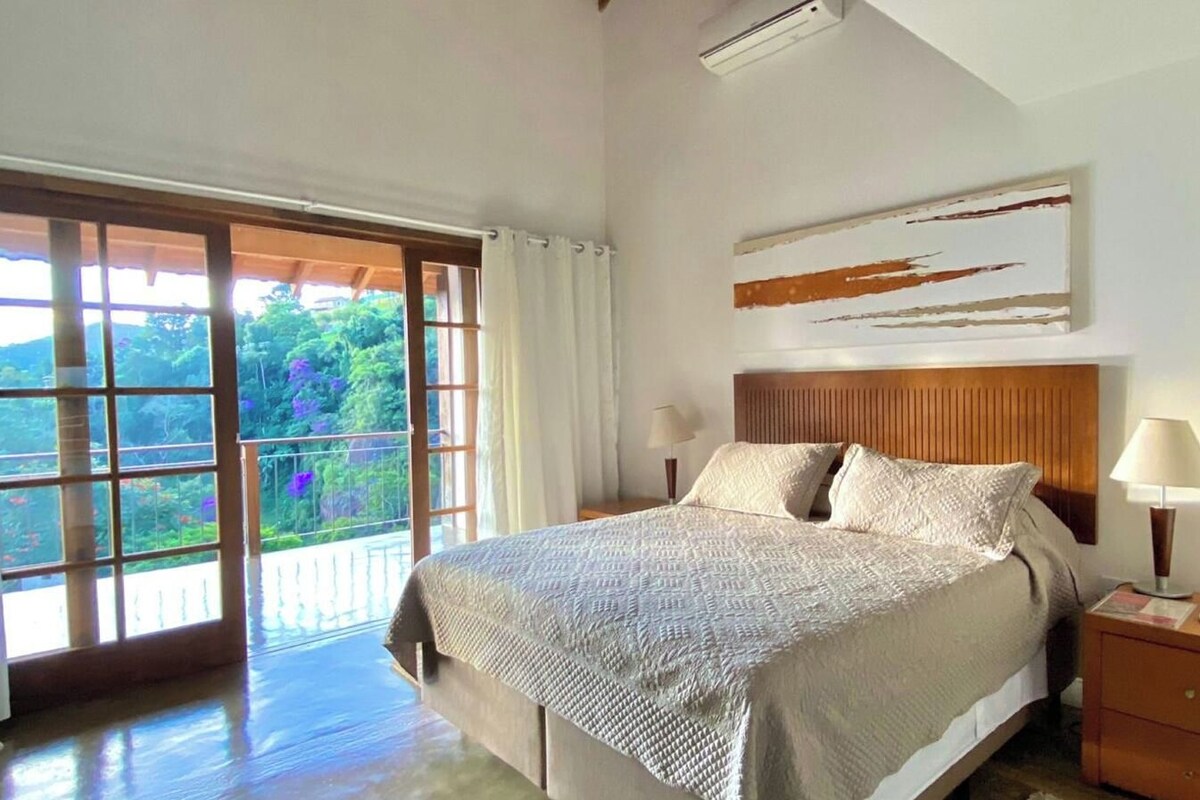 A bedroom with a large bed, a wooden headboard, a nightstand, a lamp, and a view of greenery through a sliding door.