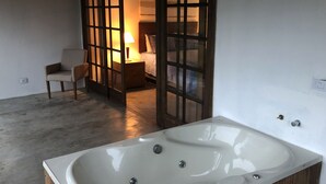 A modern bathroom with a large bathtub, a wooden cabinet, and a separate bedroom area visible through a glass door. - Pousada Golf Village (Petrópolis)